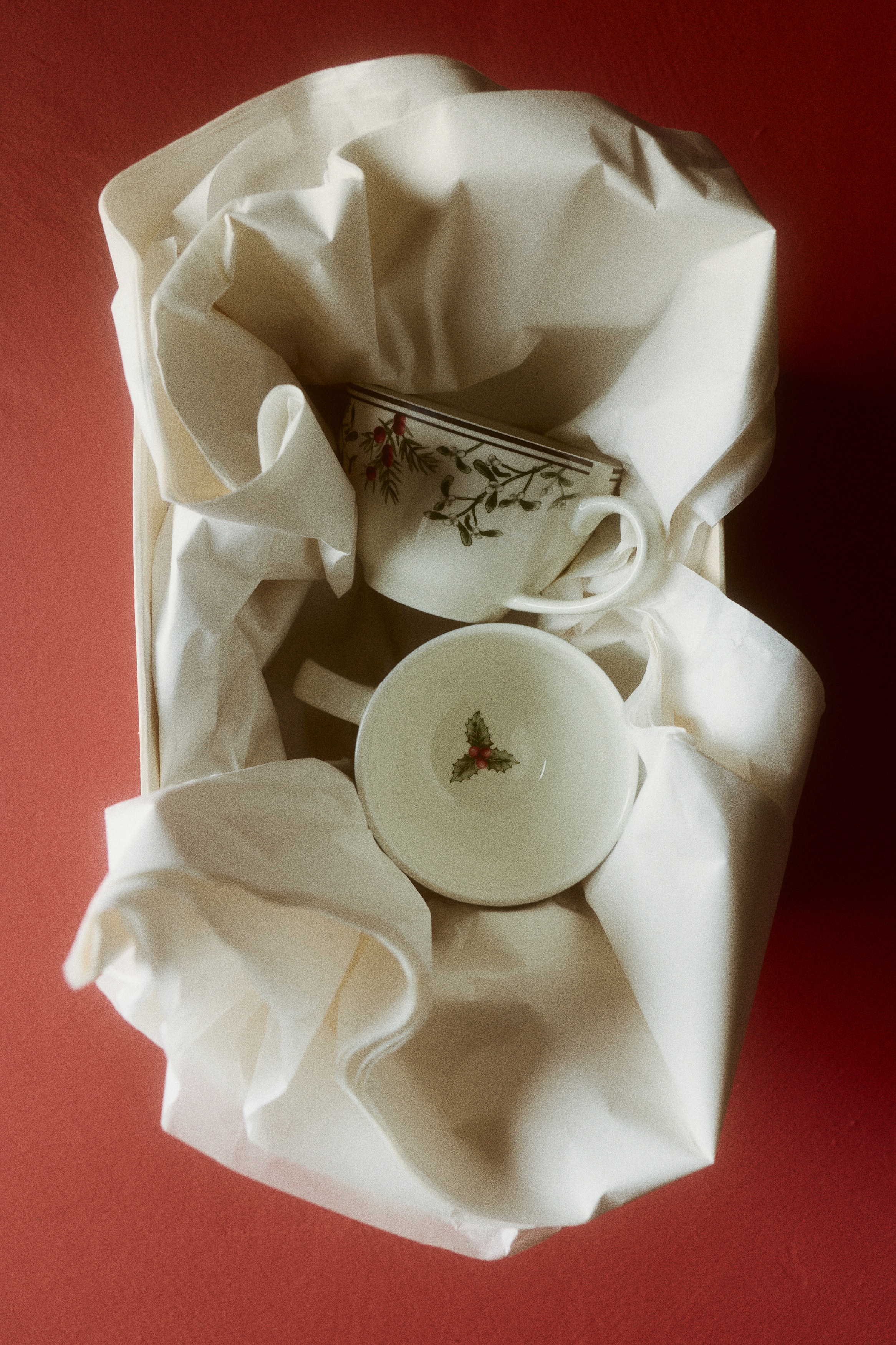 Two white ceramic teacups are sitting side by side, facing forward. Each cup displays a delicate pattern of green pine branches, holly leaves, and red berries along the upper rim, bordered by a thin dark line and featuring a curved handle on the side.
