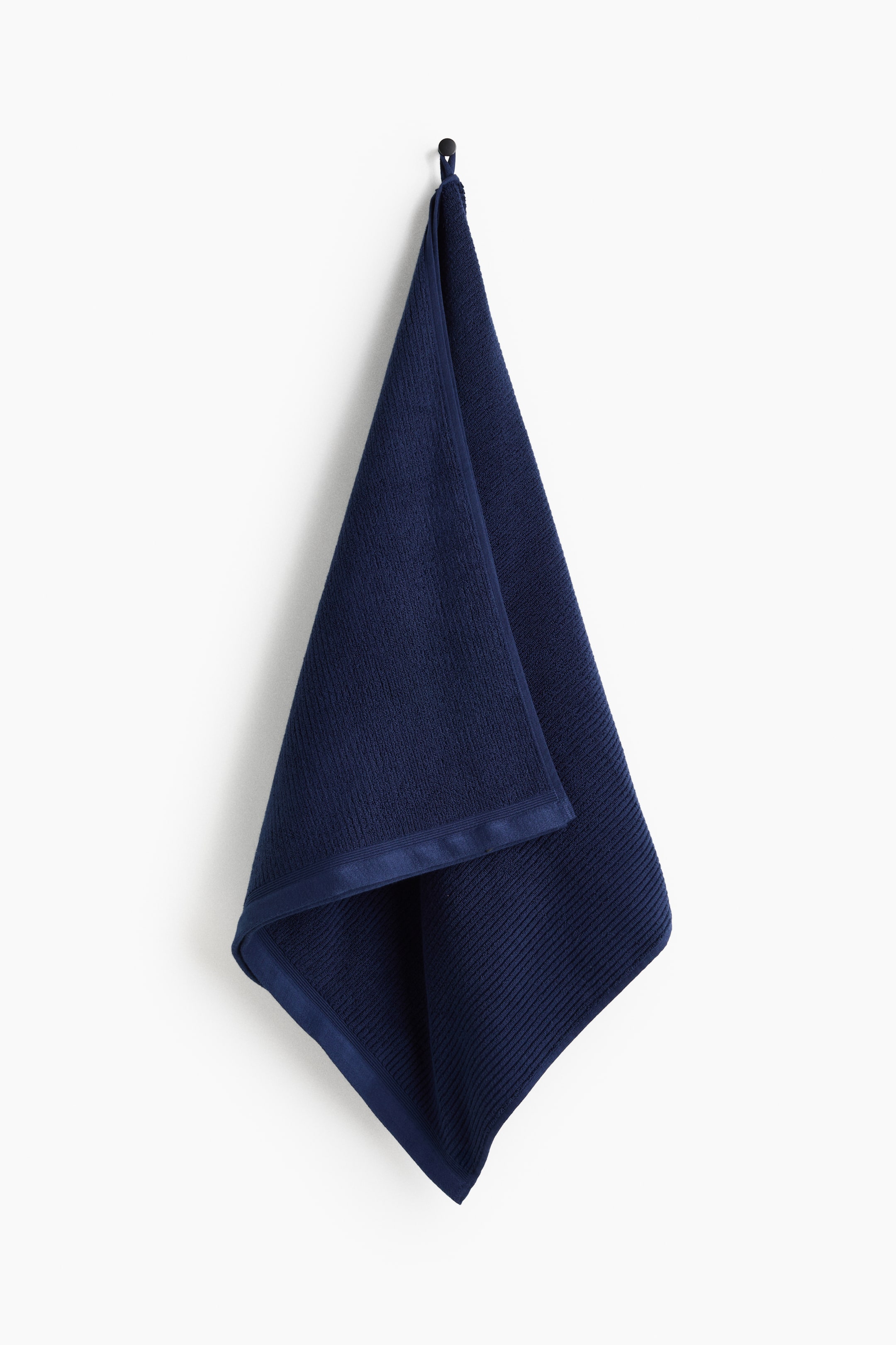 View larger image: A navy blue cotton terry bath towel is hanging from a hook by its hanger loop, displaying its textured ribbed design. The towel is draping downward, showing a smooth woven border detail along its short edges.