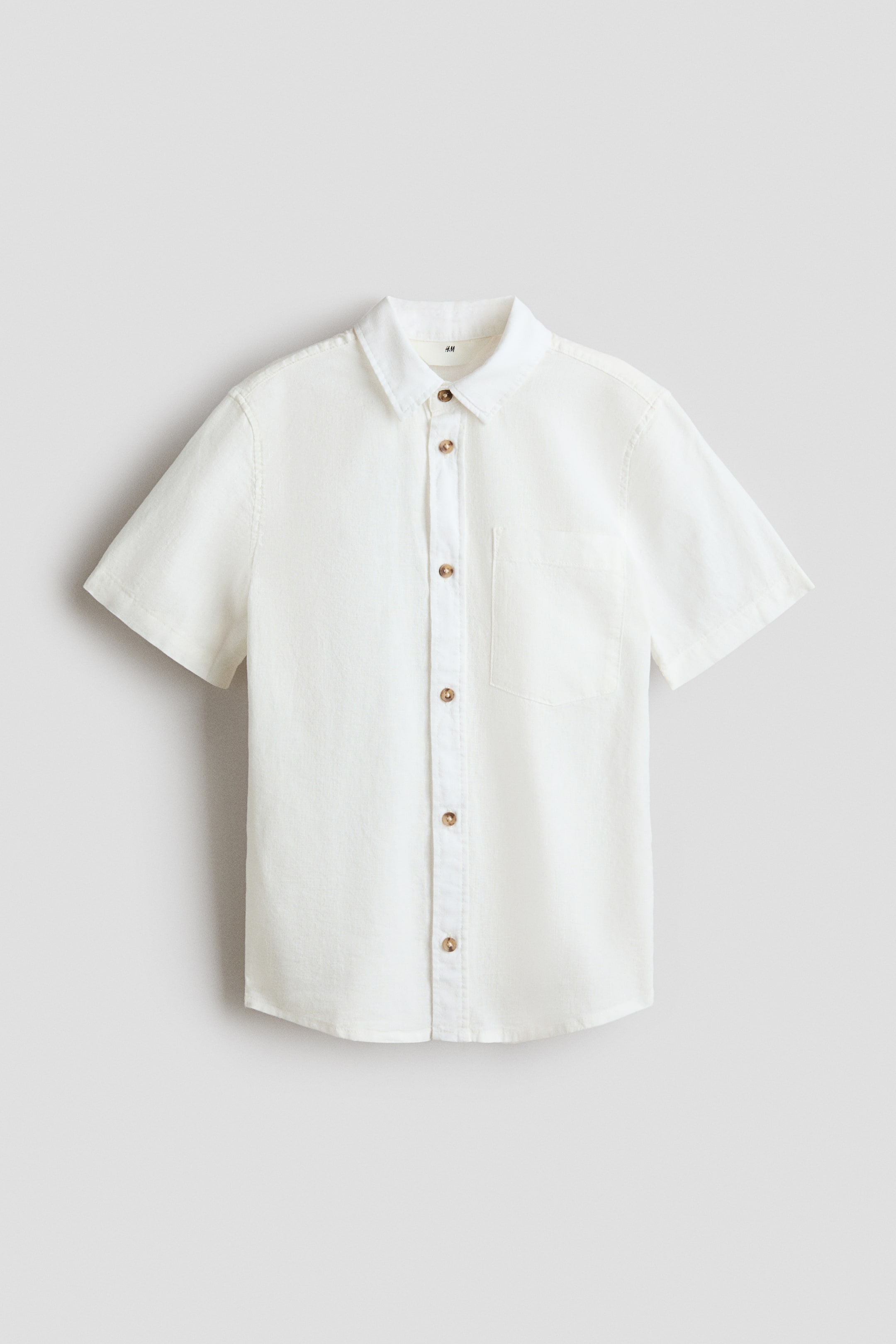 View larger image: A light-coloured, short-sleeved shirt is shown from the front, featuring textured fabric, a turn-down collar, buttons down the front, and an open left chest pocket.