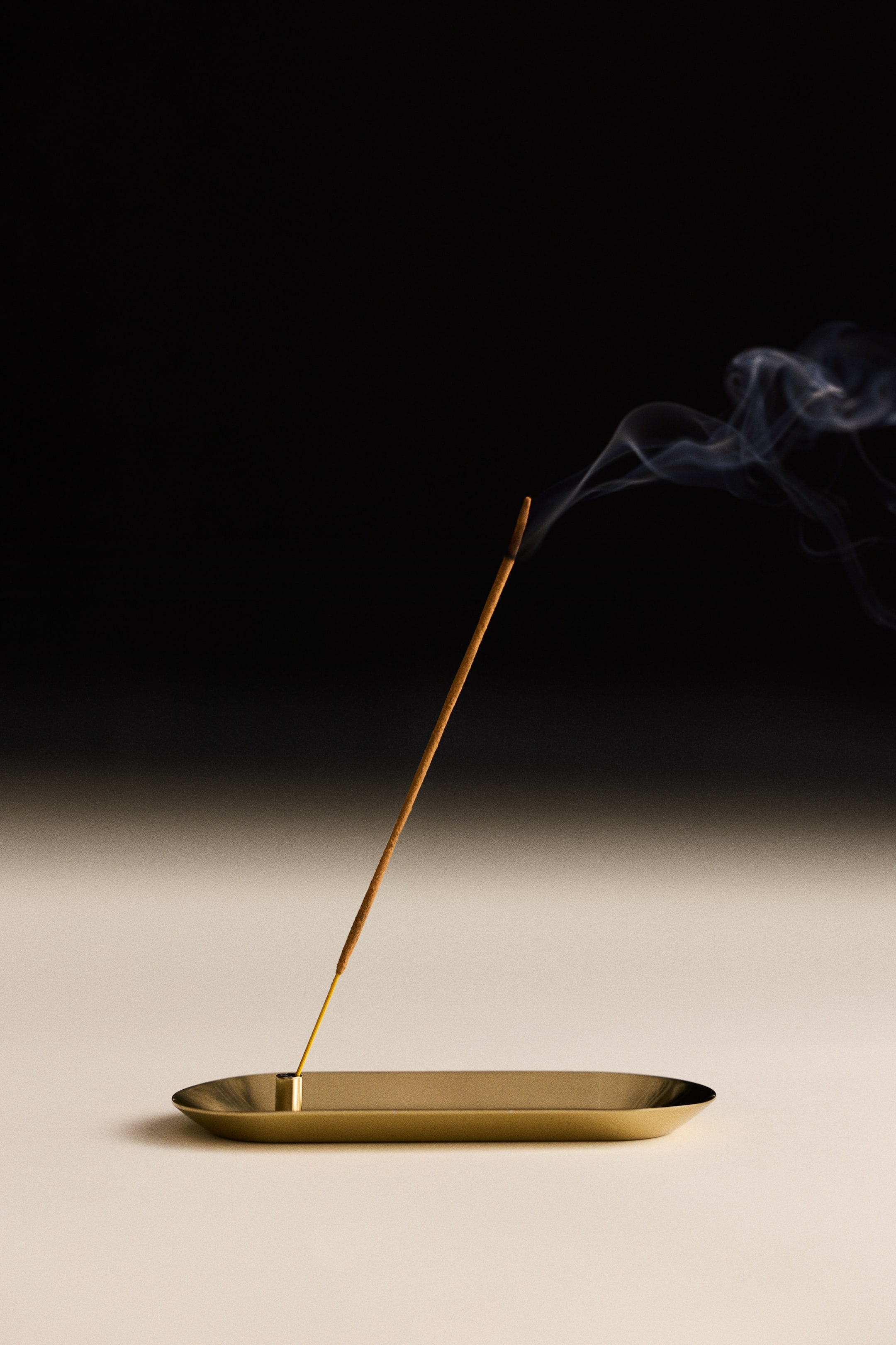 View larger image: An incense holder in gold-toned stainless steel is showcasing an oval-shaped base with a raised rim and a small cylindrical holder at one end. A single lit incense stick is resting at an angle inside the holder, releasing a thin plume of smoke.