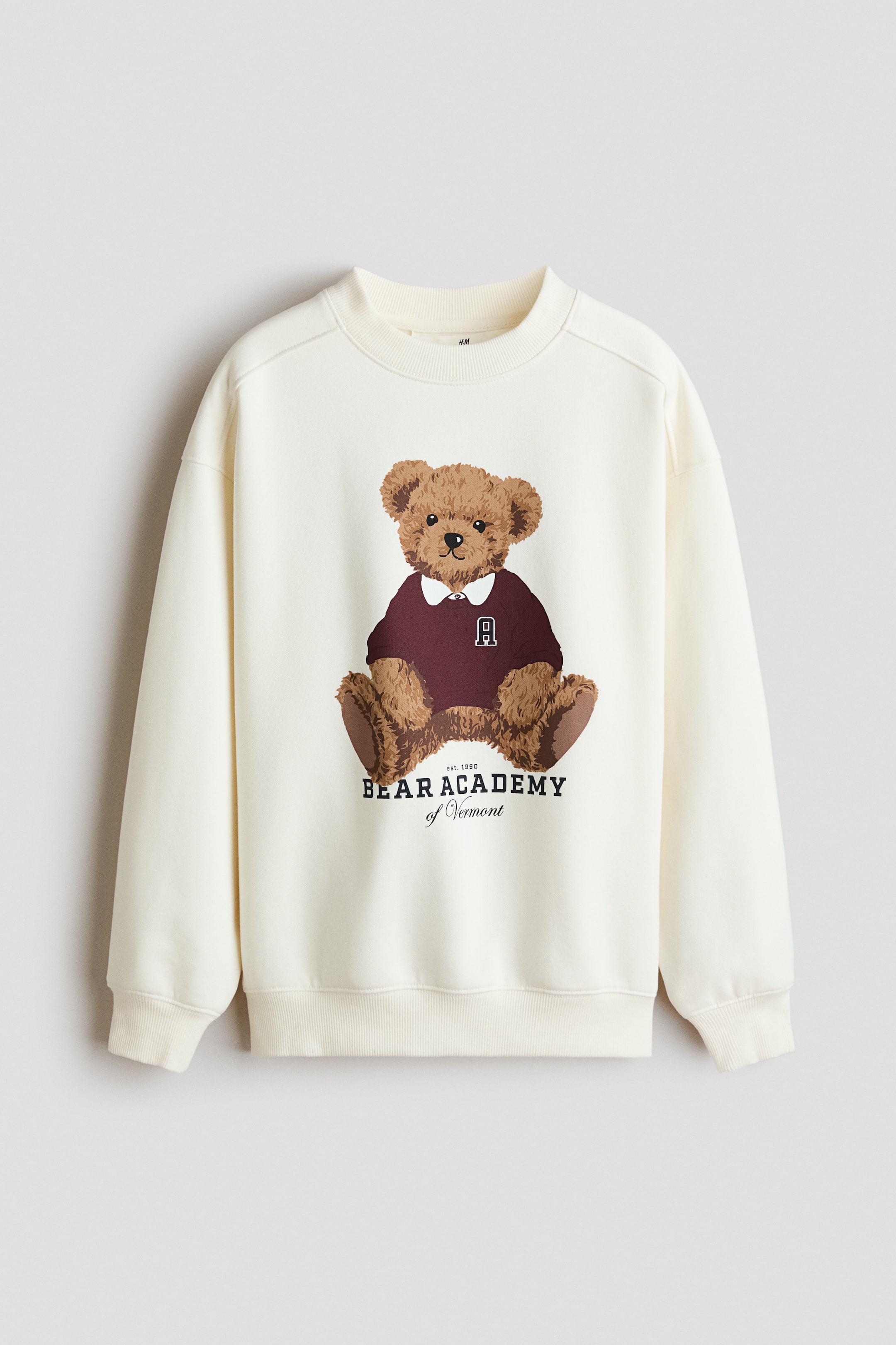Grotere afbeelding bekijken: Een crèmekleurige sweatshirt heeft een geribde ronde hals, geribde boorden en een geribde zoom en is recht van voren afgebeeld. Op de voorkant staat een grote afbeelding van een bruine teddybeer in een bordeauxrode trui met een witte kraag en een 'A'-embleem, vergezeld van de tekst "est. 1980 BEAR ACADEMY of Vermont".