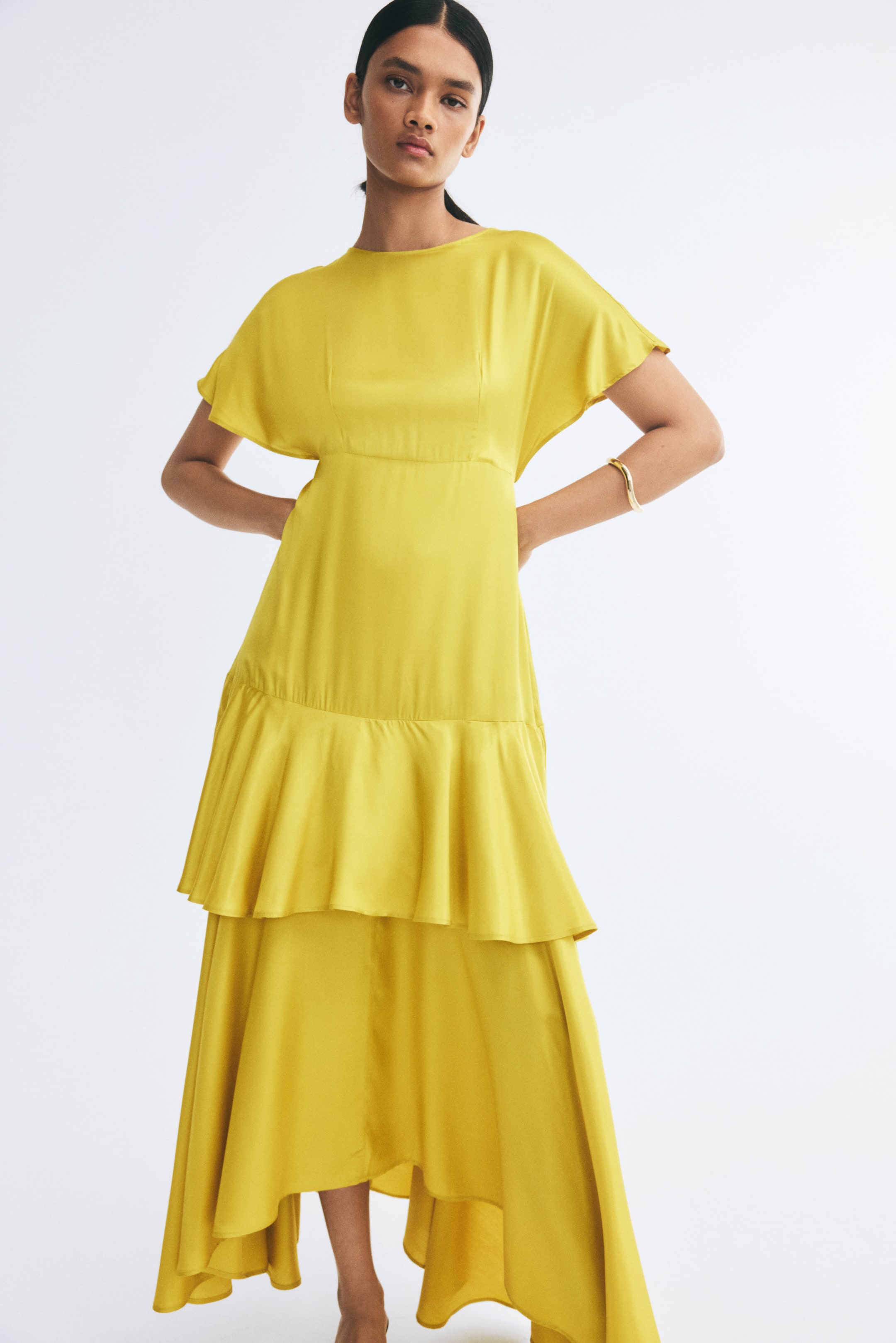View larger image: The model is wearing a long yellow dress featuring a round neckline, short butterfly sleeves, a fitted waist seam, and a tiered skirt with a flounce. She is posing with her hands placed behind her hips, highlighting the dress’s flowing high-low hem.