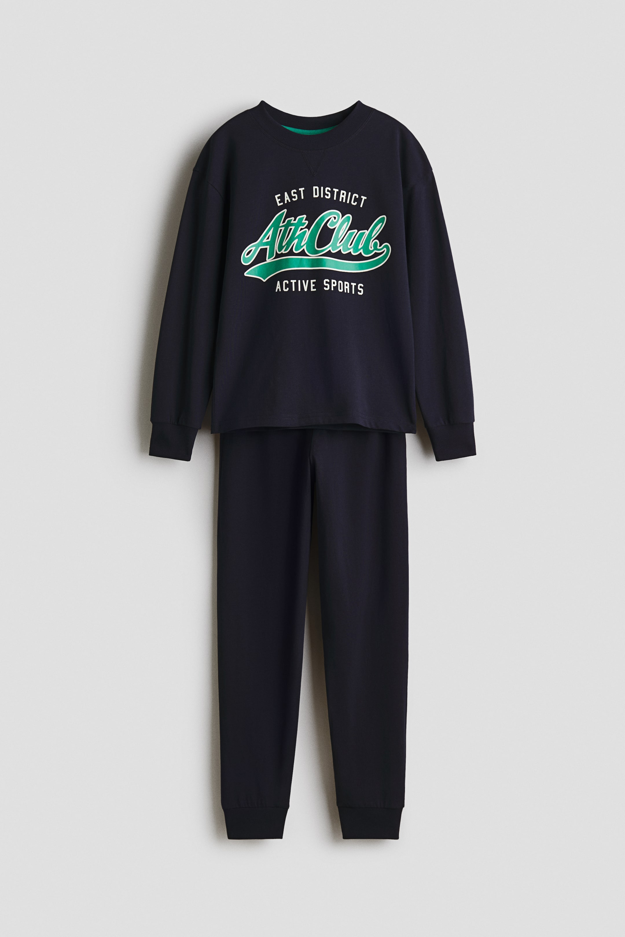 View larger image: A dark navy two-piece tracksuit is being presented, featuring a long-sleeved crew neck sweatshirt and matching jogging bottoms. The sweatshirt displays a white and green "EAST DISTRICT Air Club ACTIVE SPORTS" graphic on the chest and a green inner collar band, while both the sweatshirt and jogging bottoms have ribbed cuffs.