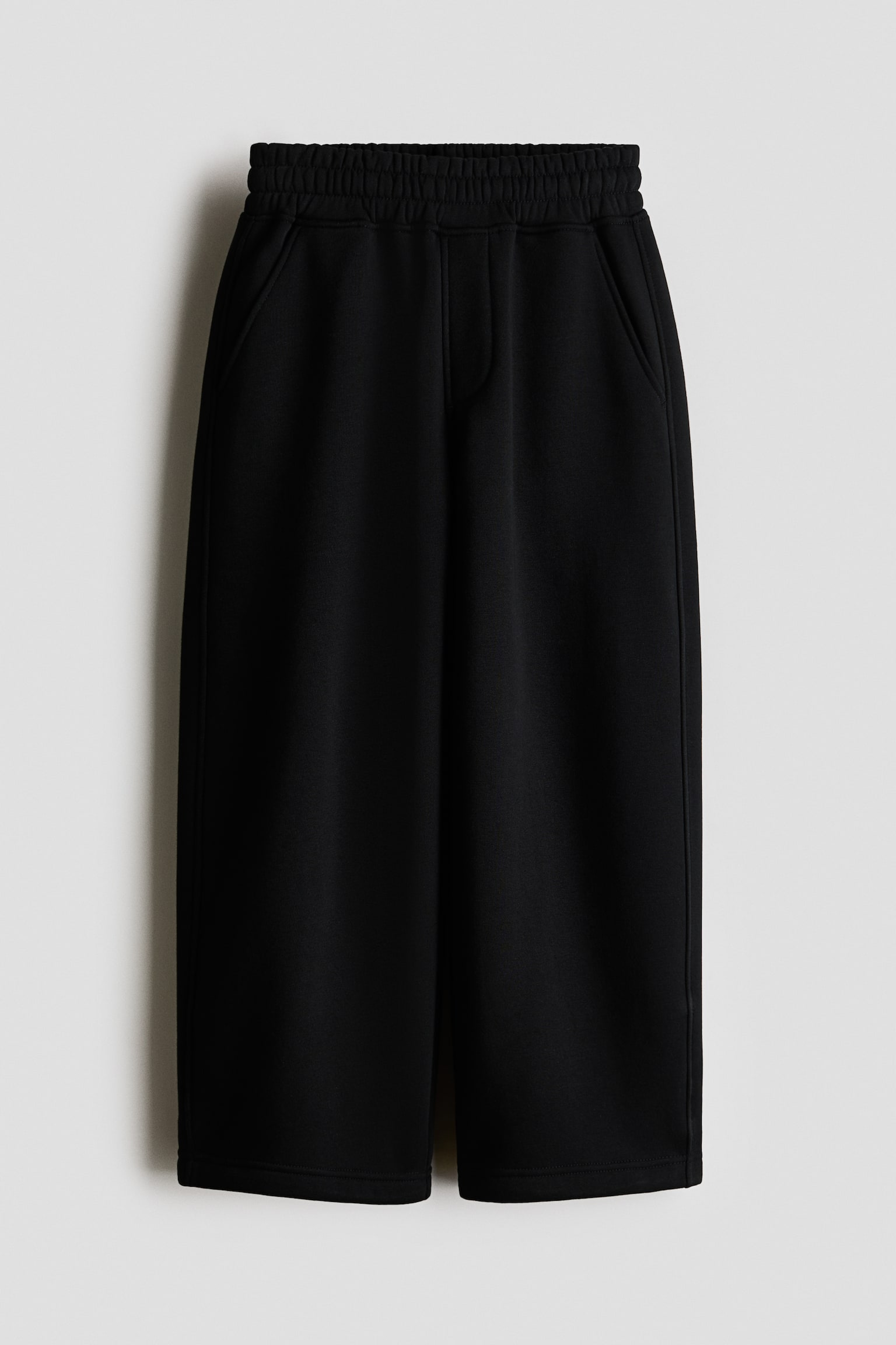 A pair of black wide-leg cropped sweatpants is hanging facing the front, featuring a thick, gathered elastic waistband, slanted side pockets, and a mock fly detail. The pants are displaying a relaxed, loose fit with wide, straight-cut legs. - 1