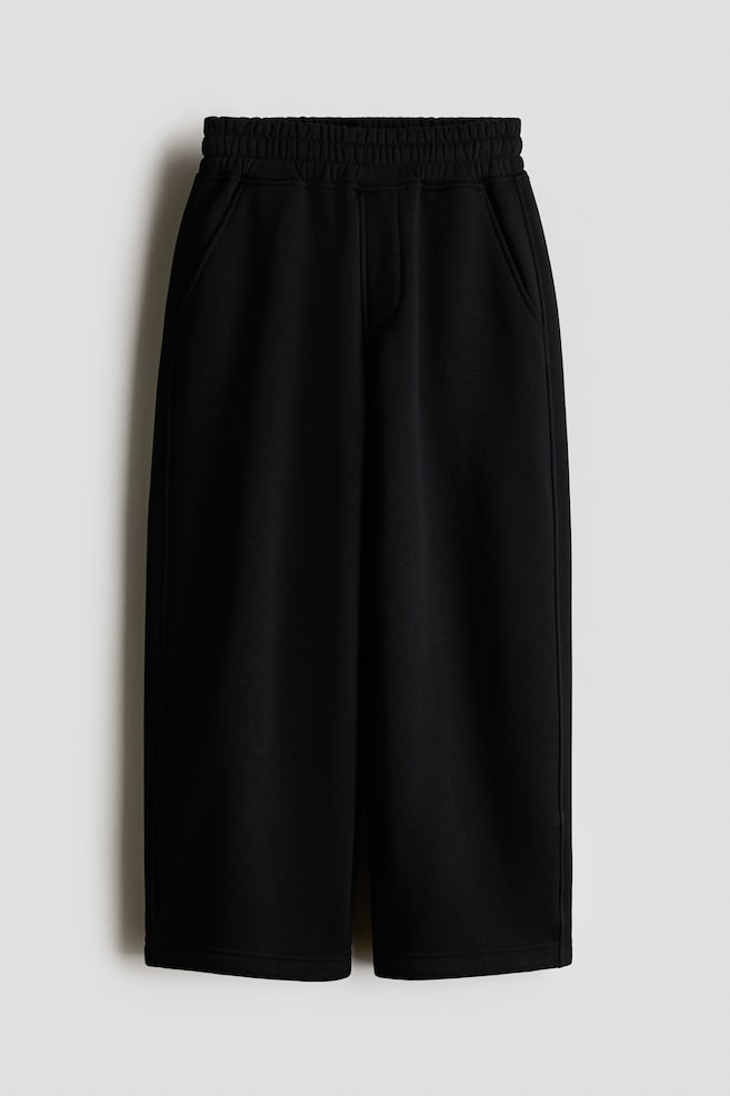 A pair of black wide-leg cropped sweatpants is hanging facing the front, featuring a thick, gathered elastic waistband, slanted side pockets, and a mock fly detail. The pants are displaying a relaxed, loose fit with wide, straight-cut legs. - 1