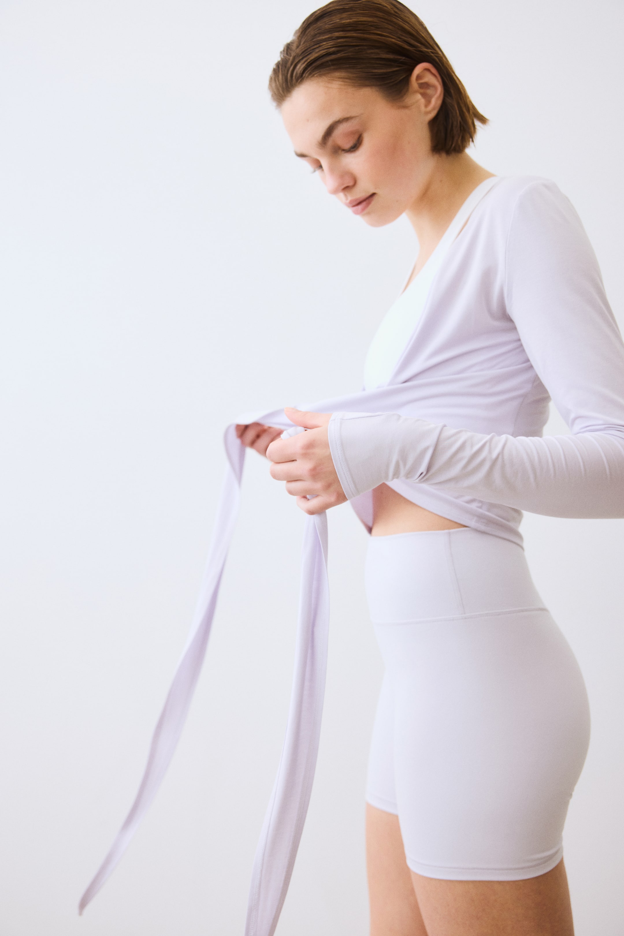 View larger image: A light purple long-sleeve wrap top is tied at the front, showcasing an overlapping V-neck and extended cuffs. It is worn over a white sports bra and paired with matching high-waisted athletic shorts that reach mid-thigh.
