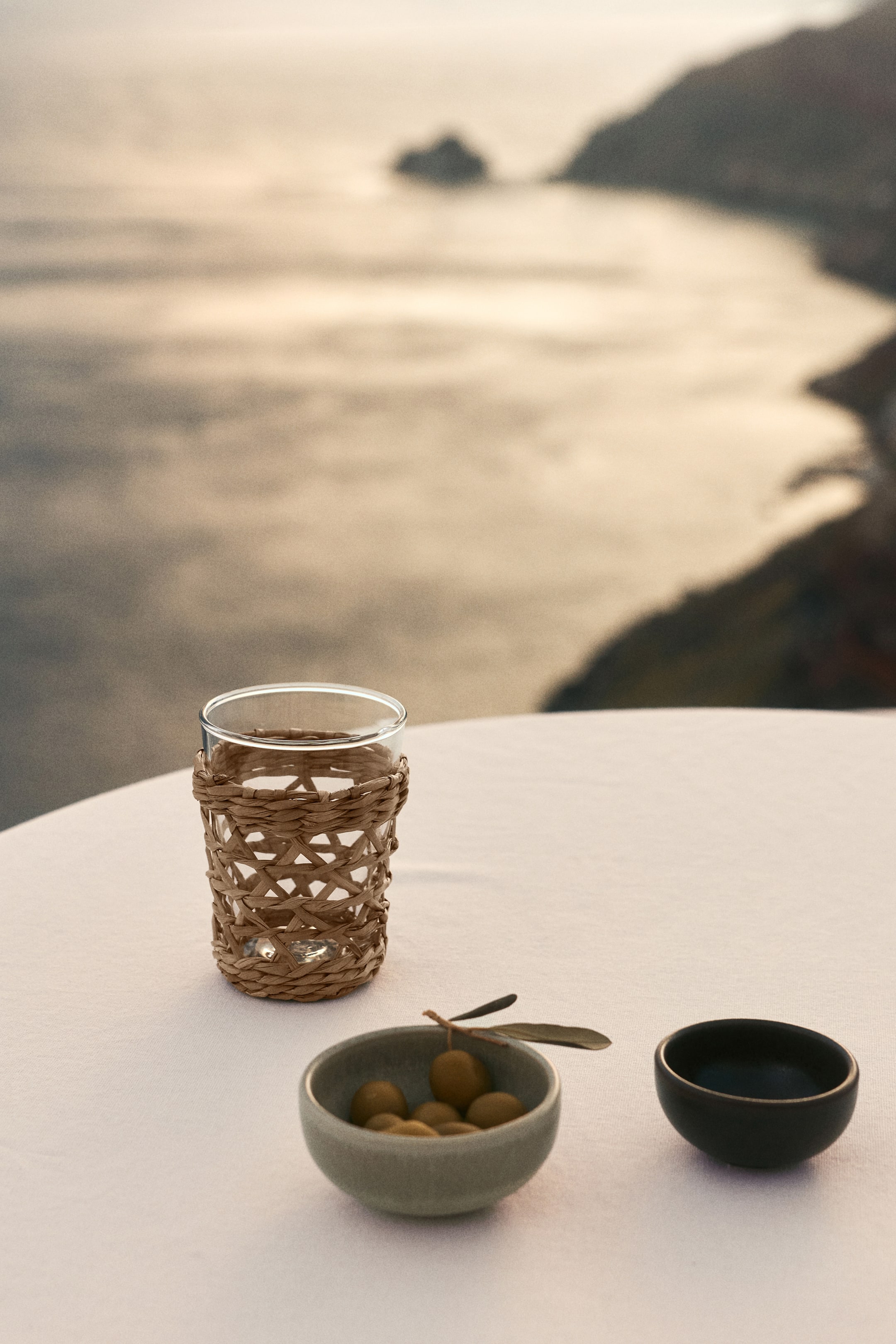 View larger image: A transparent glass, adorned with a woven straw holder showcasing a crisscross design around its lower part, is placed on a white tablecloth. Beside it, there's a light green ceramic bowl filled with green olives and an olive branch, accompanied by a small, dark ceramic bowl featuring an angled rim.