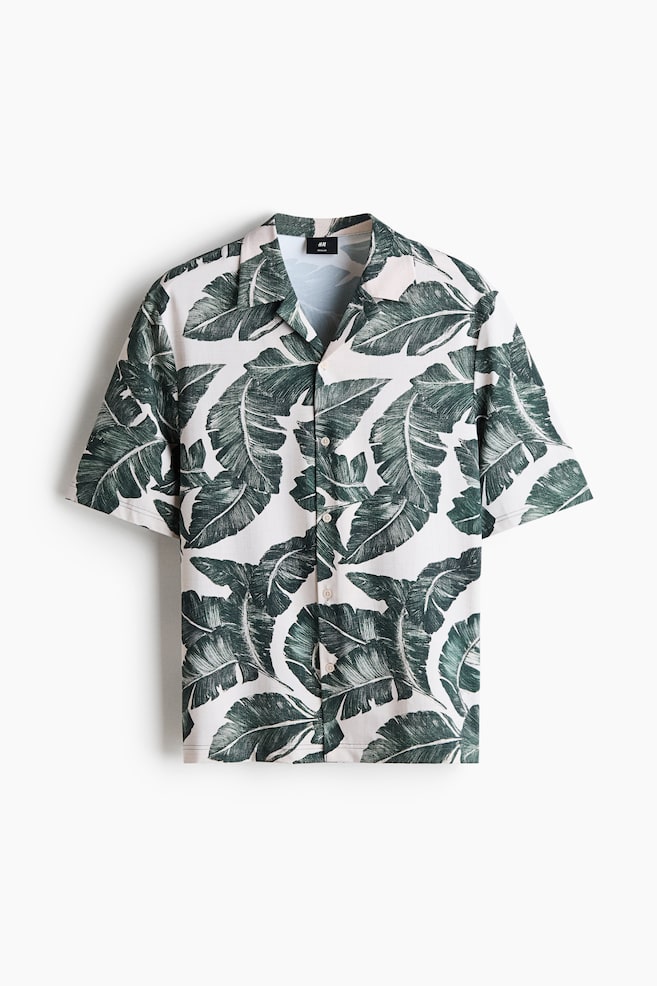 A short-sleeved shirt showcases a white background adorned with a prominent dark green banana leaf print. It features a camp collar and a full button-front closure with light-coloured buttons. - 1
