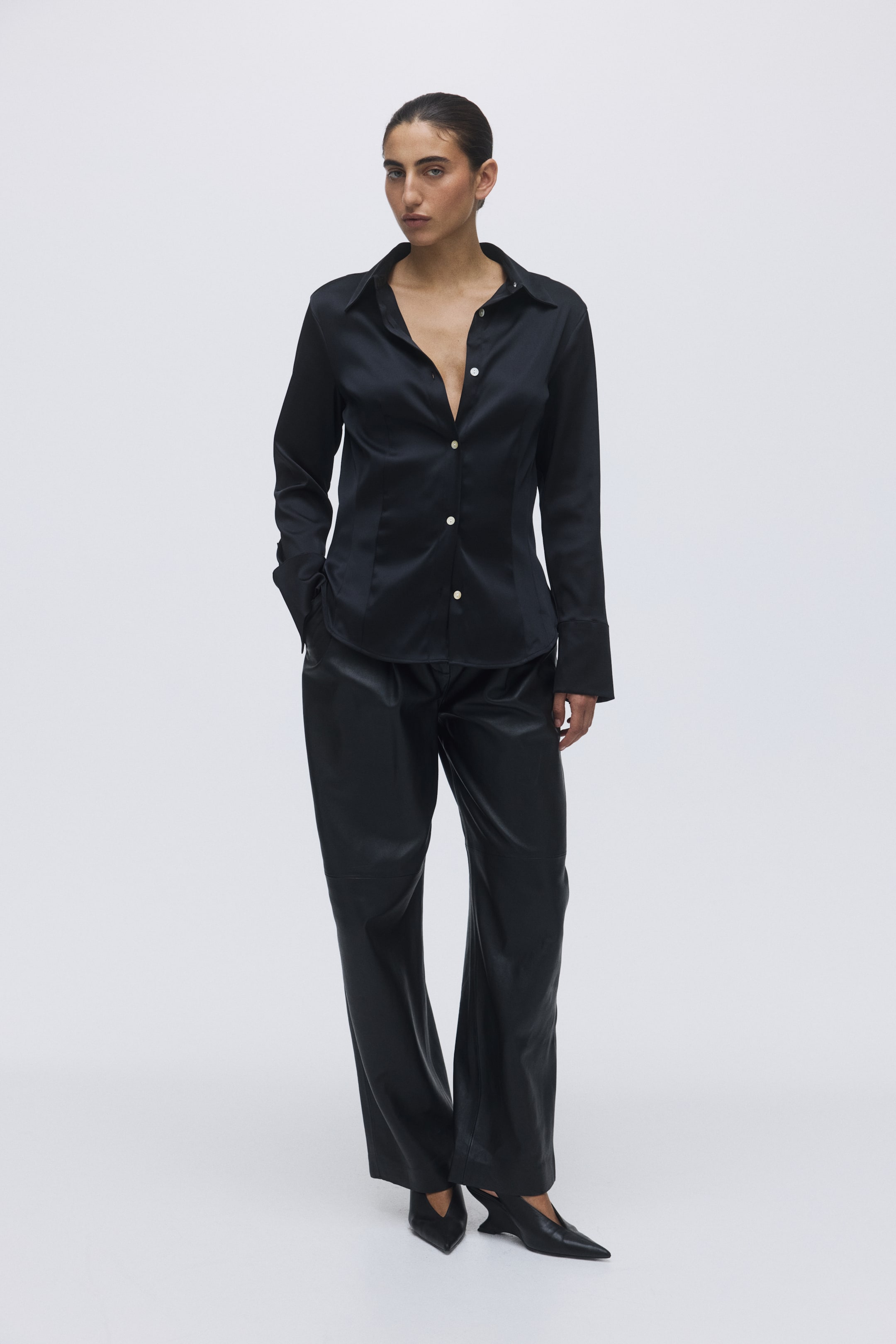 이미지 확대 보기: 검은색 광택이 나는 긴 소매 셔츠는 뾰족한 칼라와 단추가 달린 커프스를 특징으로 하며, 검은색 와이드 팬츠와 함께 착용되고, 뾰족한 발가락의 검은색 신발로 스타일링되어 있습니다. 셔츠는 부분적으로 단추가 풀려 V자형 네크라인을 만들고 있으며, 바지는 여유로운 핏으로 전체적인 앙상블을 정면에서 보여줍니다.