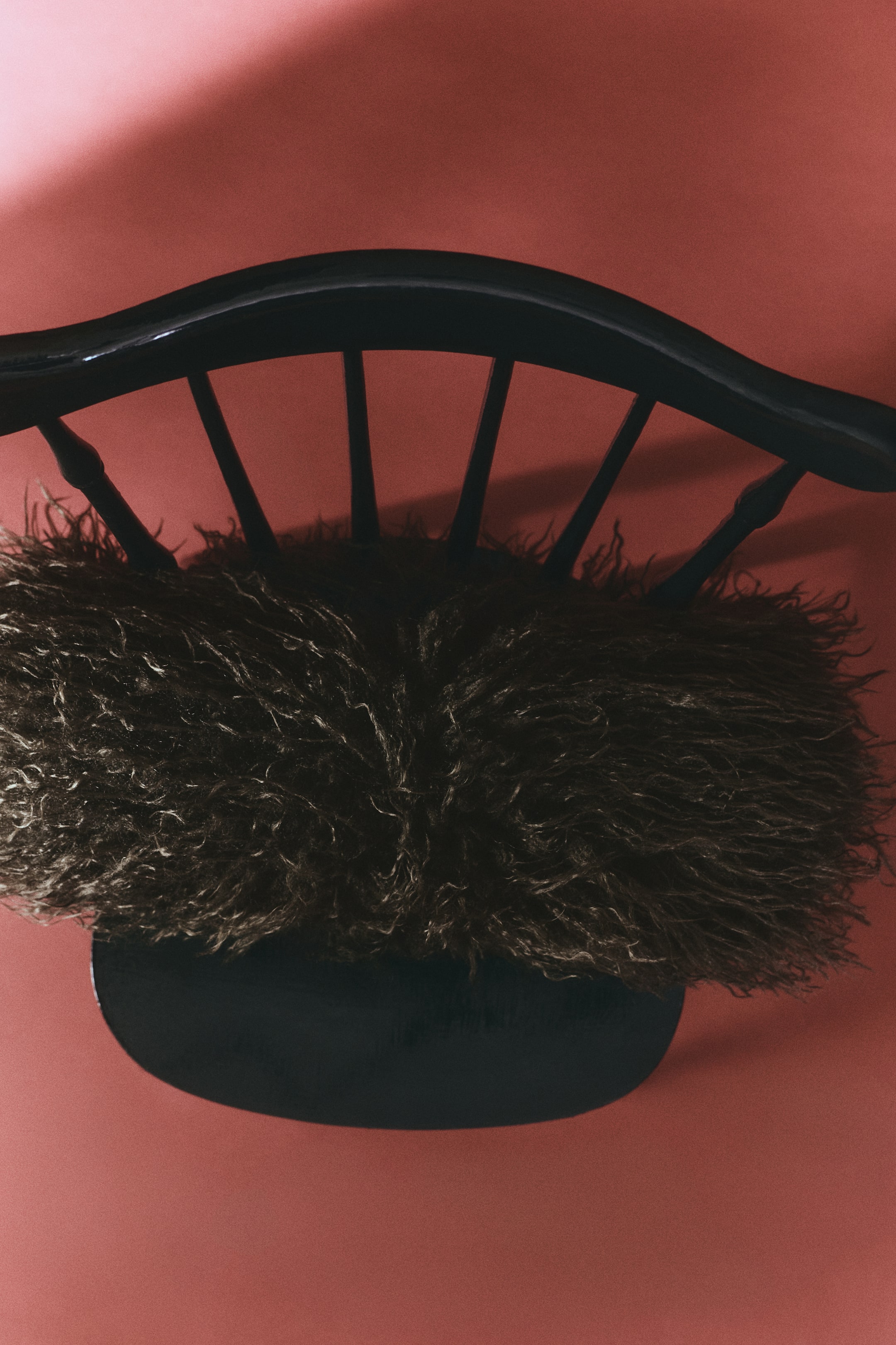 View larger image: A bolster cushion covered in dark brown, long-pile fluffy fabric is resting horizontally on the seat of a black wooden spindle-back chair, displaying its dense, shaggy texture.