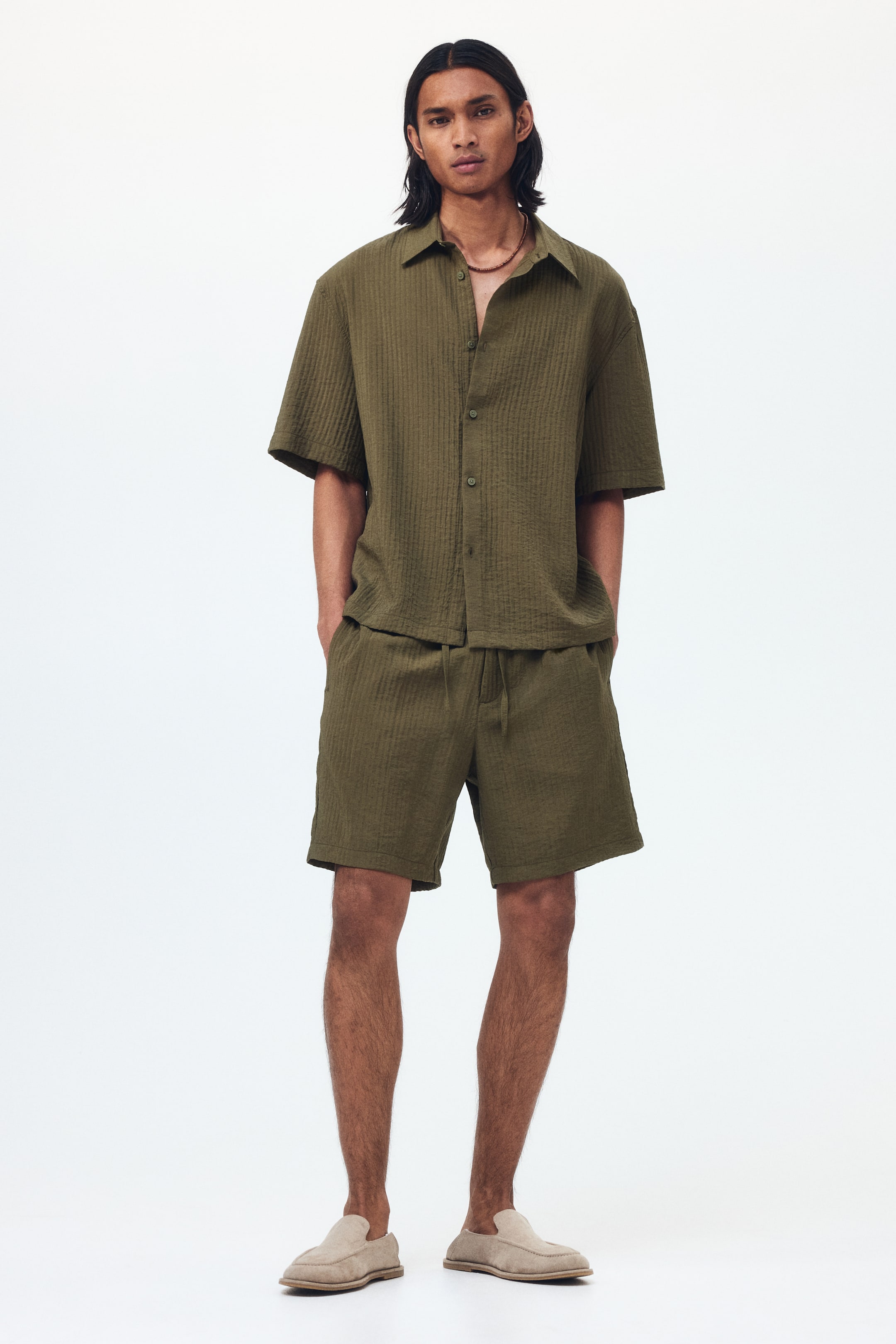 Visualizza l'immagine ingrandita: Un completo composto da una camicia a maniche corte verde oliva e pantaloncini coordinati viene mostrato in vista frontale. La camicia presenta un colletto classico e una chiusura frontale con bottoni, mentre i pantaloncini sono progettati con un girovita con coulisse, entrambi i capi esibendo un tessuto increspato e strutturato.