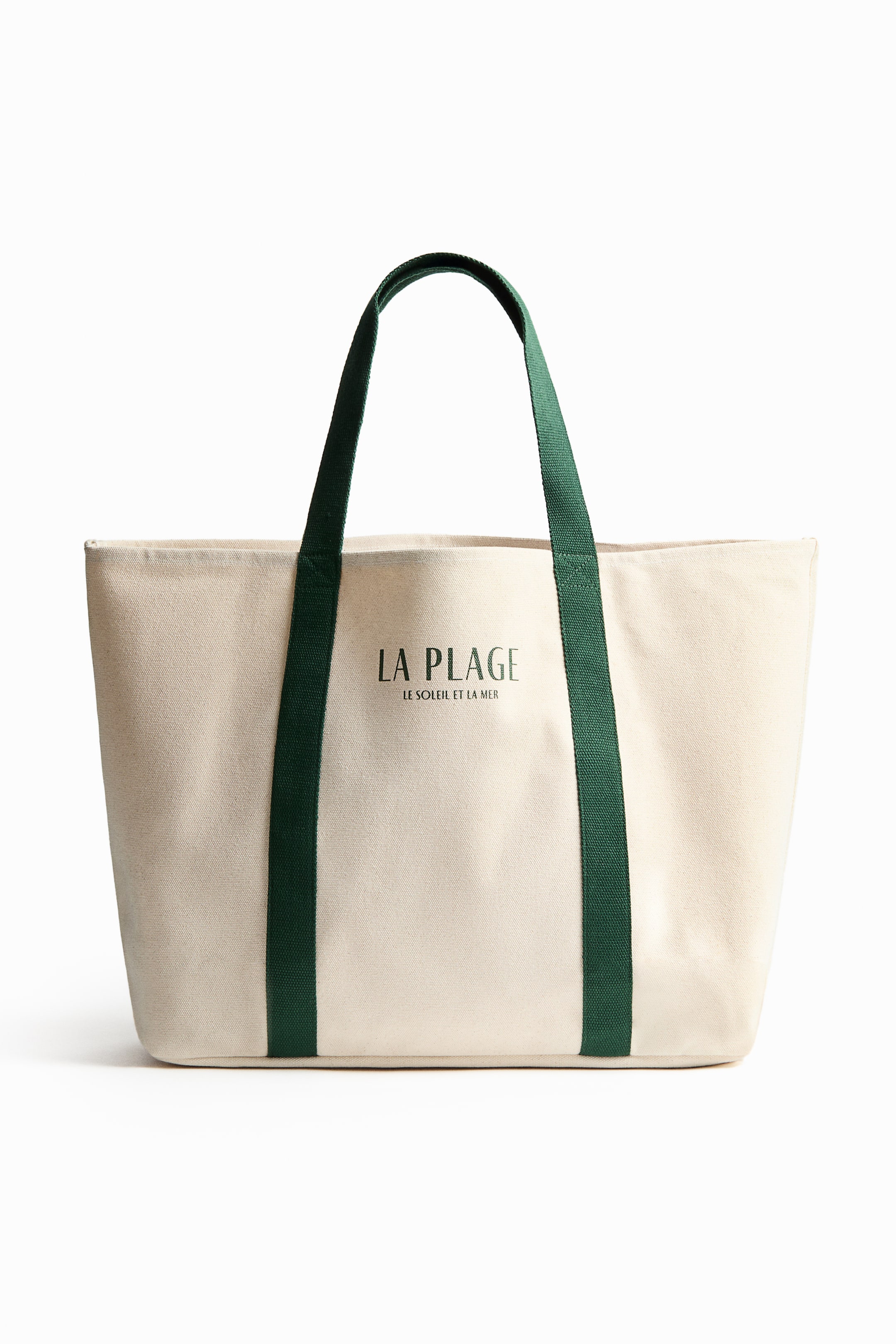 拡大画像を表示: ビーチバッグは、頑丈なコットンキャンバスで作られた正面を紹介しています。上部にはアクセントカラーのハンドルが付いており、正面には「LA PLAGE LE SOLEIL ET LA MER」と書かれたプリントモチーフがあります。
