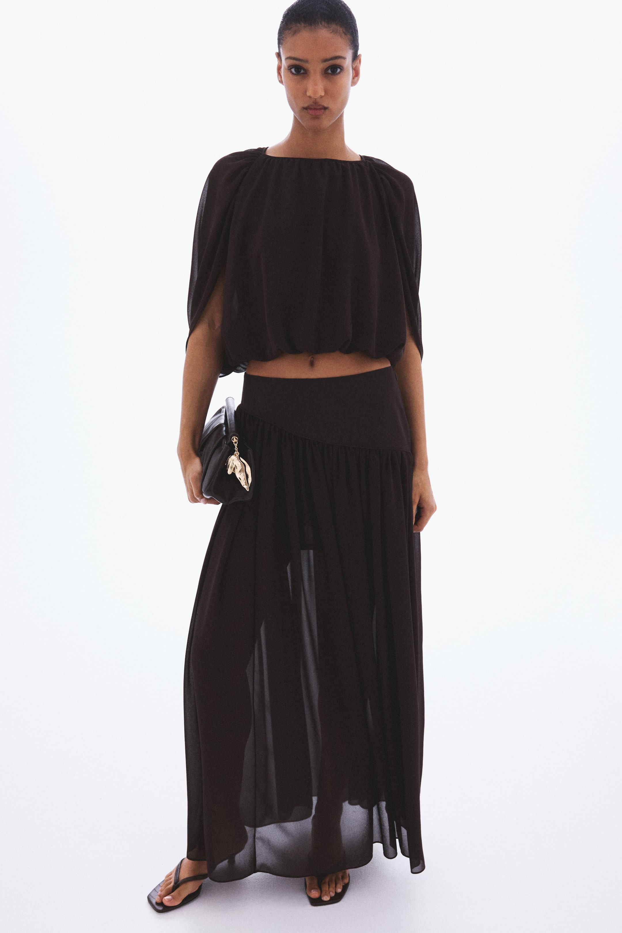 View larger image: The product is a dark two-piece set, featuring a cropped blouson top and a sheer maxi skirt. The top presents a gathered boat neckline, wide short sleeves, and an elasticized hem. The skirt showcases an elasticized high waist, a full flowing silhouette, and an opaque lining visible through the sheer outer fabric.