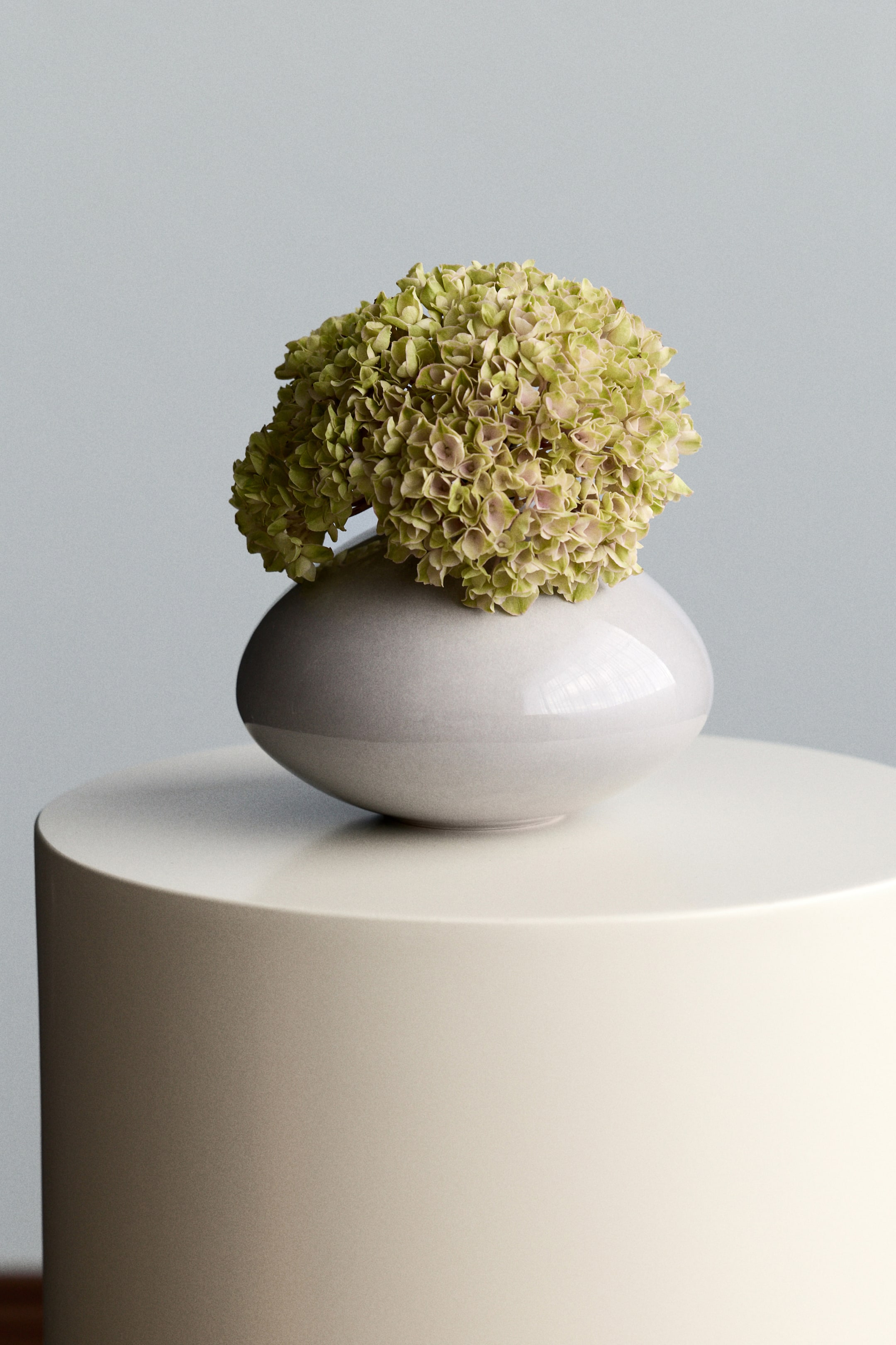 View larger image: A light grey, glossy ceramic vase is showcasing a cluster of greenish-yellow hydrangeas, its rounded form resting on a cream-coloured cylindrical stand.