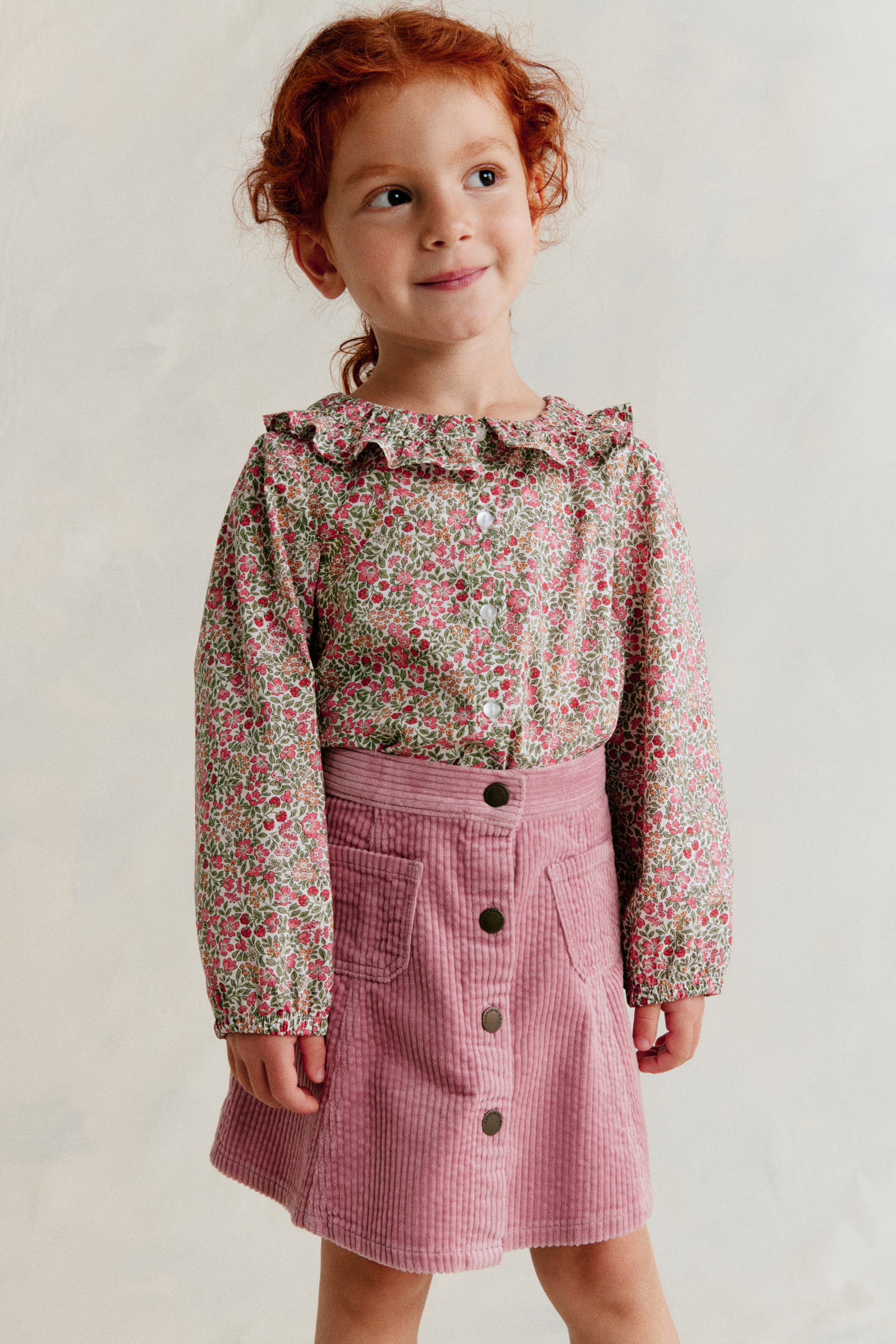View larger image: The blouse is facing front, showcasing a sweet floral print with pink flowers and green leaves. A frill collar adorns the neckline, white buttons run down the front, and long, voluminous sleeves gather at the wrists with elasticated cuffs.