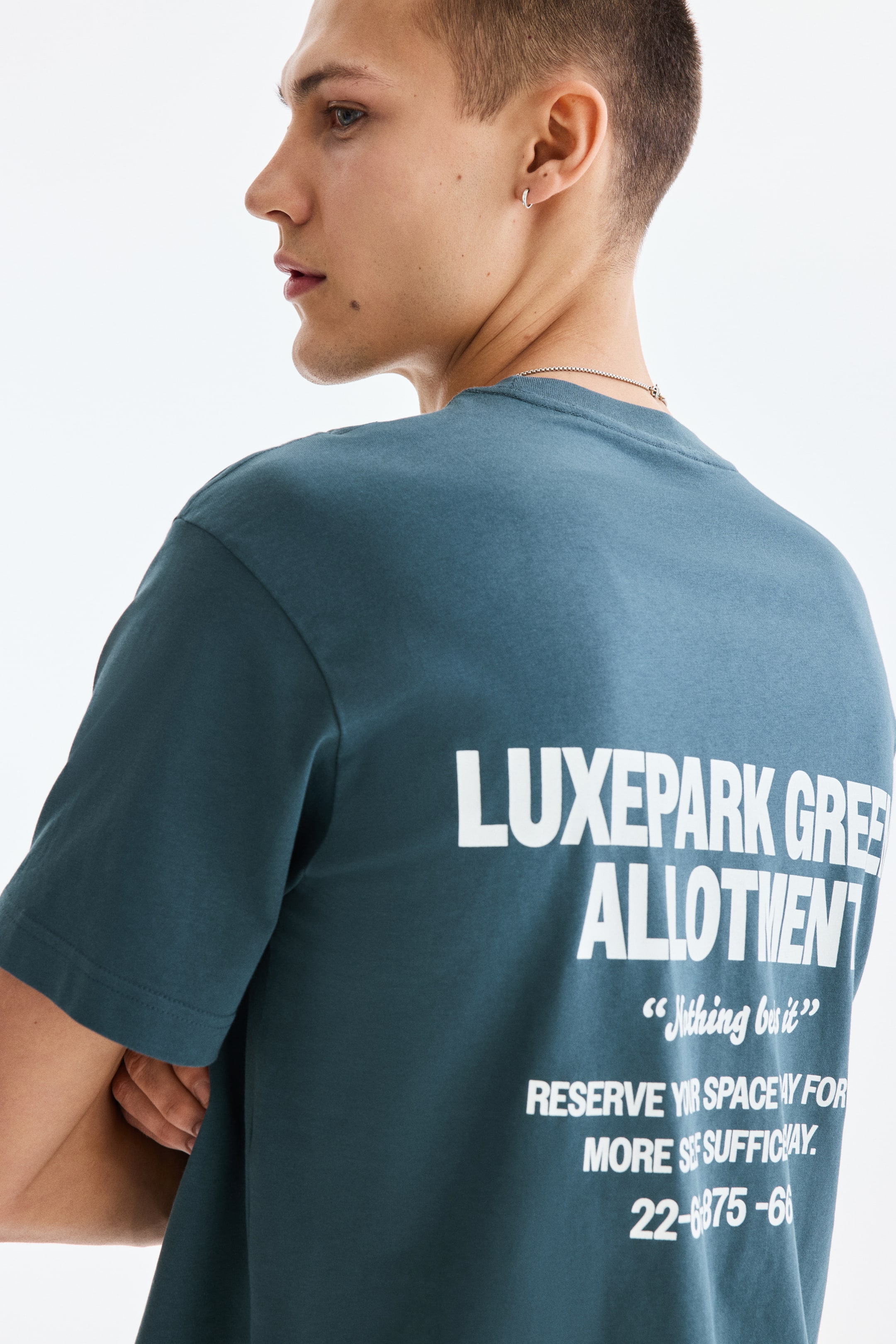 View larger image: A dark teal t-shirt is worn from the back, highlighting its rounded crew neck and short sleeves. The back of the t-shirt showcases a large white graphic print, featuring prominent text that reads "LUXEPARK GRE ALLOTMENT" with additional lines of smaller text beneath it.