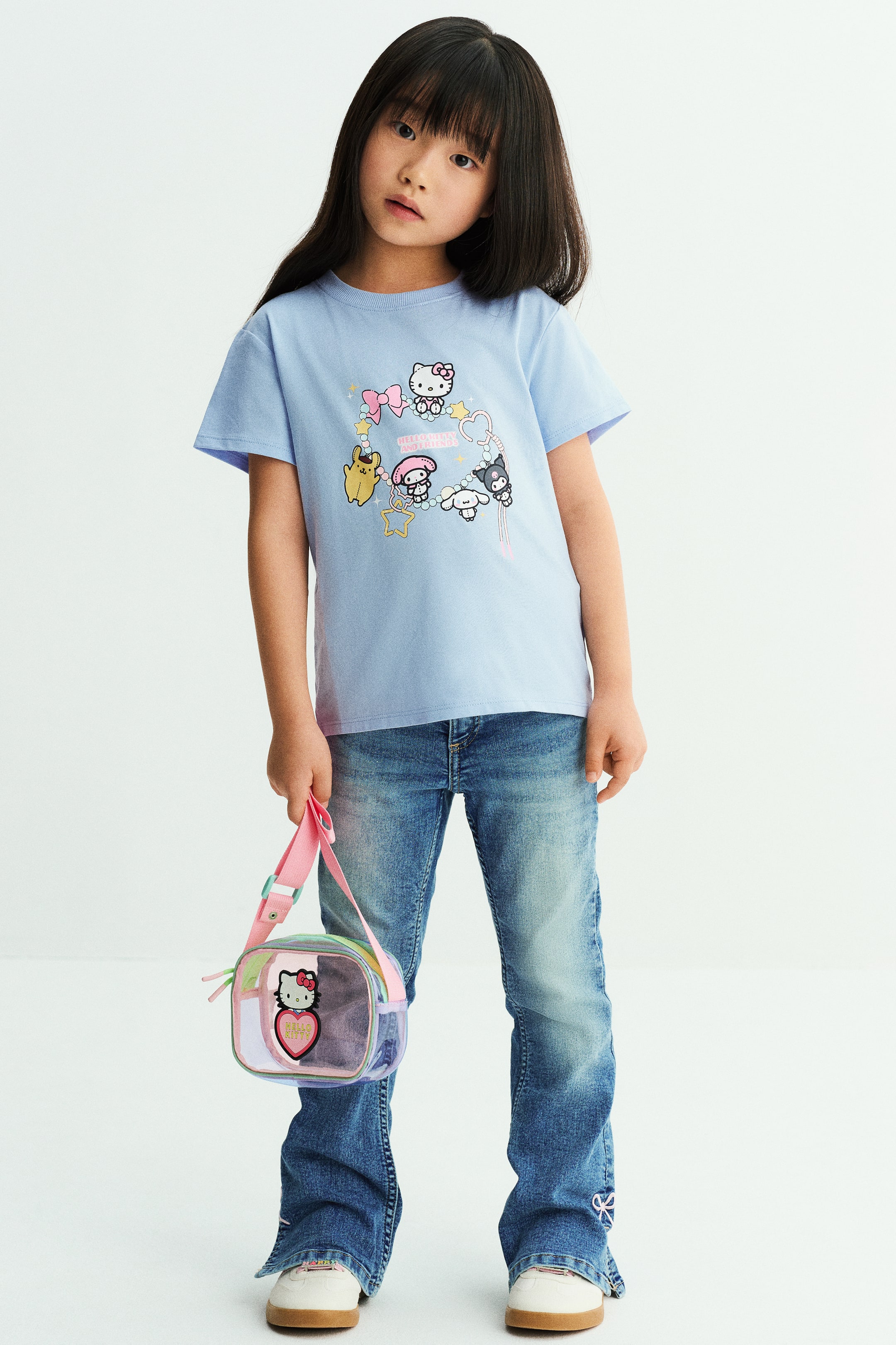 View larger image: A light blue cotton jersey T-shirt featuring a colourful cartoon print on the front, a round rib-trimmed neckline, and dropped shoulders is worn by the model. The model styles the T-shirt with blue jeans while holding a small, transparent handbag with a pink strap.