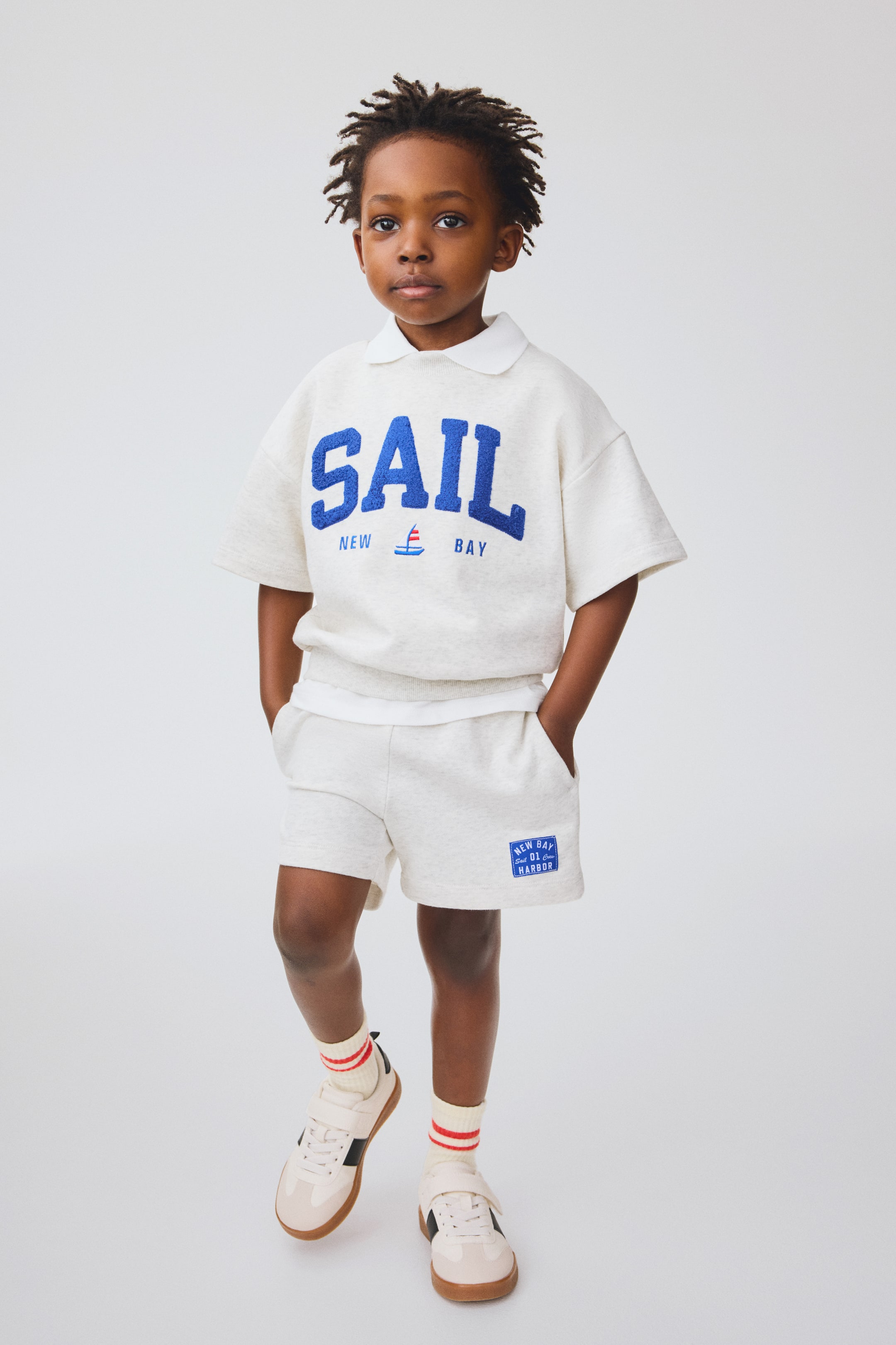 View larger image: A light gray marl short-sleeved top is showcased, featuring a white polo collar and blue chenille 'SAIL' lettering across the chest, with 'NEW,' a sailboat graphic, and 'BAY' in smaller text below. The matching shorts feature side pockets and a blue badge on the left leg that reads 'NEW CO. 01 HARBOR'.