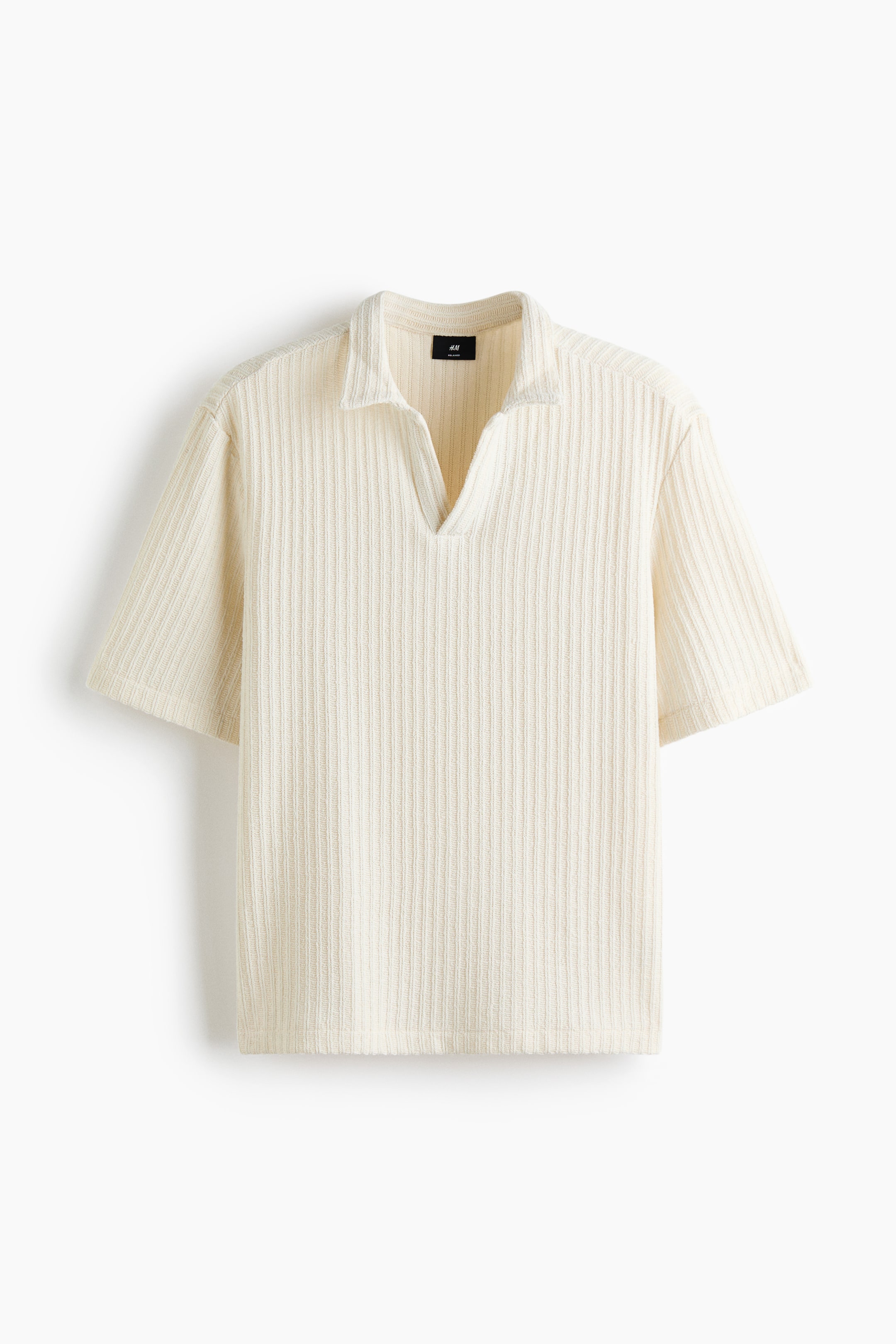 Visualizza immagine più grande: Una polo a maniche corte color crema è distesa in piano, mettendo in risalto una lavorazione a maglia jacquard a coste verticali. Presenta un colletto e un'apertura a forma di V al collo, offrendo una silhouette rilassata.
