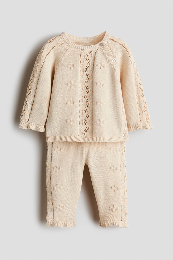 Un ensemble de tricot pour bÃ©bÃ© de couleur crÃ¨me est prÃ©sentÃ© de face, composÃ© dâun pull Ã  manches longues et dâun pantalon assorti, ornÃ©s de motifs floraux en tricot texturÃ©. Le pull prÃ©sente une fermeture asymÃ©trique Ã  boutons le long de la couture raglan, des dÃ©tails en pointelle et des poignets volantÃ©s, tandis que le pantalon affiche des panneaux latÃ©raux en pointelle assortis et des ourlets volantÃ©s. - 1