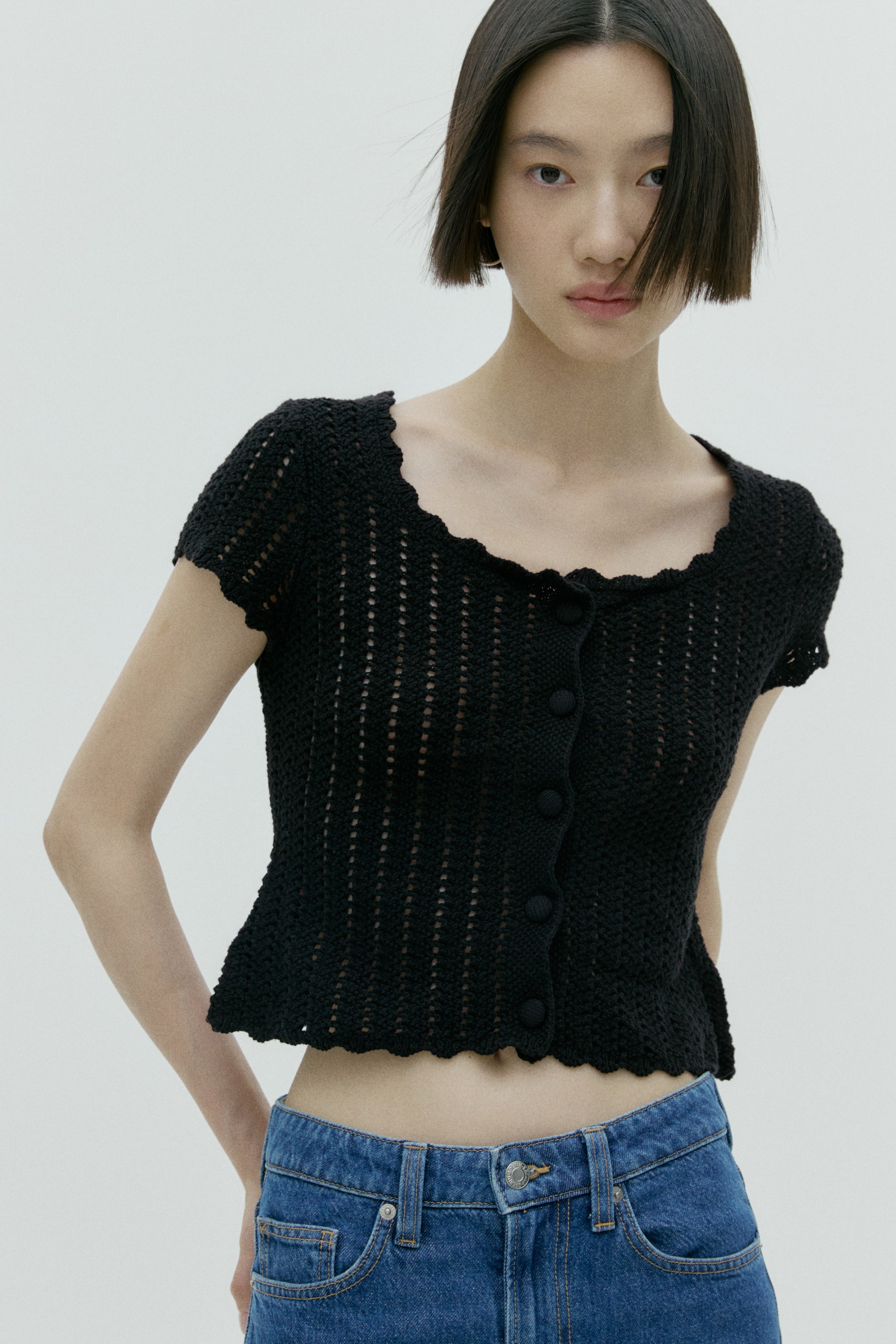 View larger image: A black crocheted short-sleeve top features an open-knit pattern, a scalloped square neckline, and a button-front closure with multiple black buttons. The hem gently flares and has a scalloped edge.