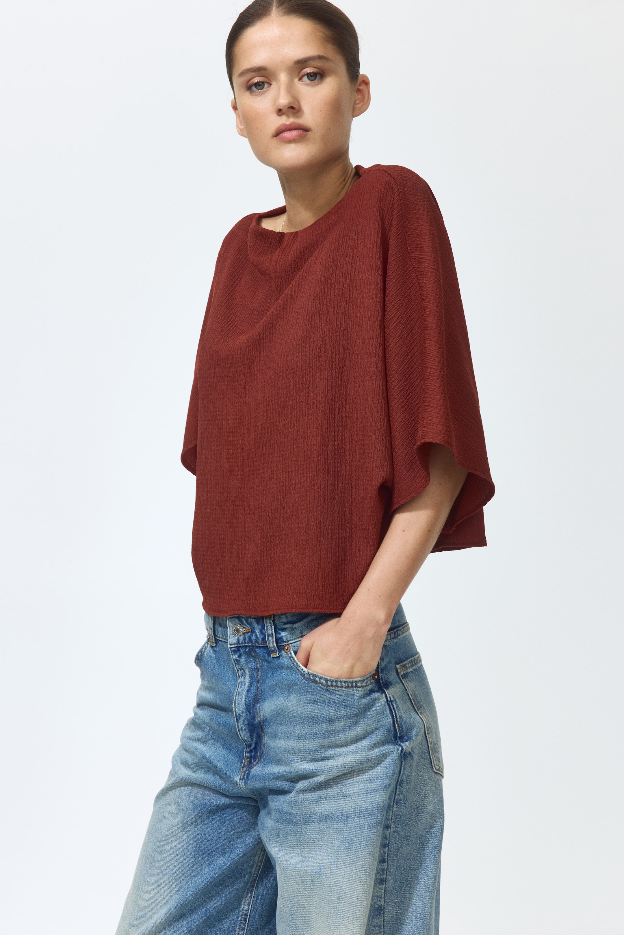 Grotere afbeelding bekijken: Een roestkleurige, boxy-stijl top in gekreukelde jersey, met een verfijnde boothals, ellebooglange vleermuismouwen en een recht gesneden zoom, wordt gedragen door het model.