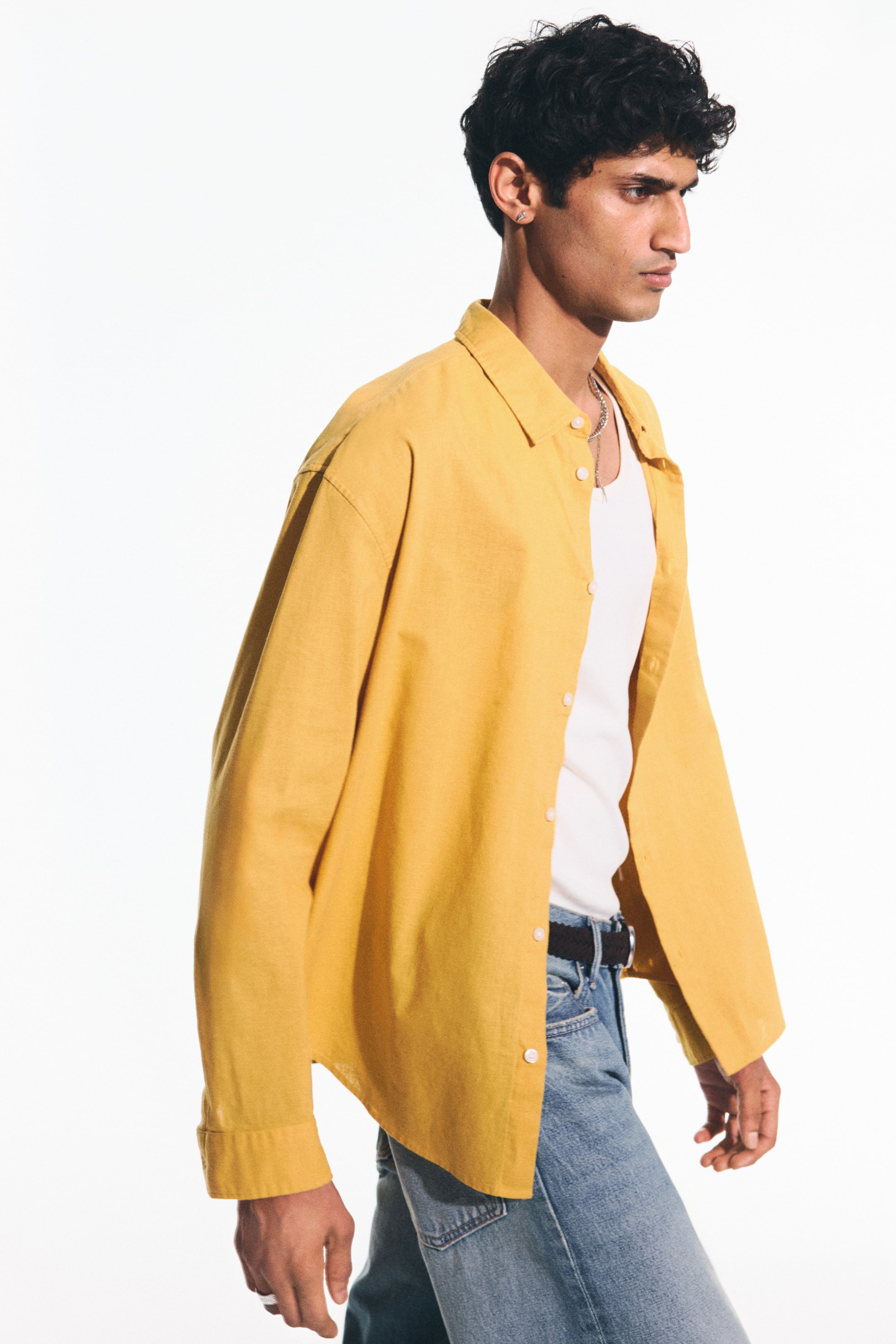 View larger image: Presenting a yellow button-up shirt from a side profile, highlighting its relaxed fit. It features a pointed collar, long sleeves with cuffs, and white buttons down the front, styled open over a white tank top.