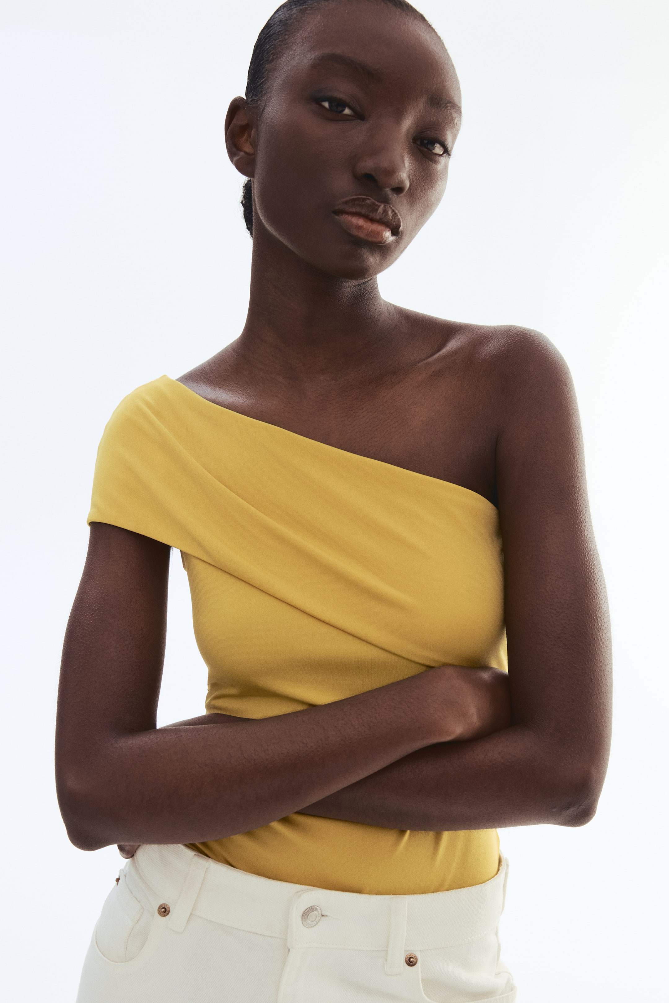 View larger image: The model is wearing a mustard yellow one-shoulder bodysuit crafted from stretch jersey. It showcases a foldover design at the top and is paired with light-colored pants.