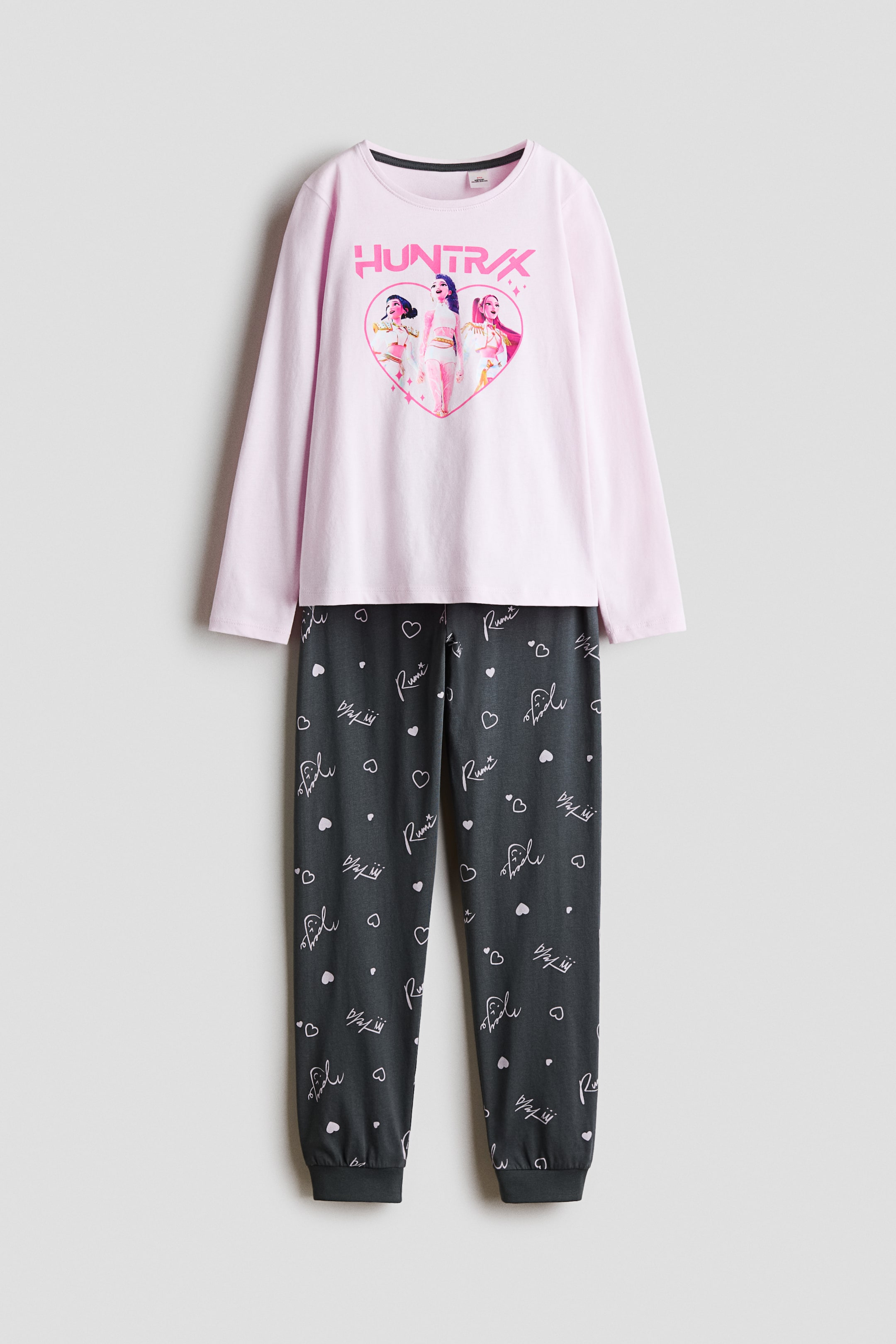 Größeres Bild ansehen: Ein vorne dargestelltes Pyjama-Set mit einem hellrosa, langärmeligen Oberteil mit Rundhalsausschnitt und einem Front-Grafikprint mit drei Figuren in Herzform unter dem Wort „HUNTRX“. Die dazugehörige dunkelgraue Hose zeigt ein Allover-Muster aus Herzen und Schriftzügen und ist mit gerippten Knöchelbündchen versehen.