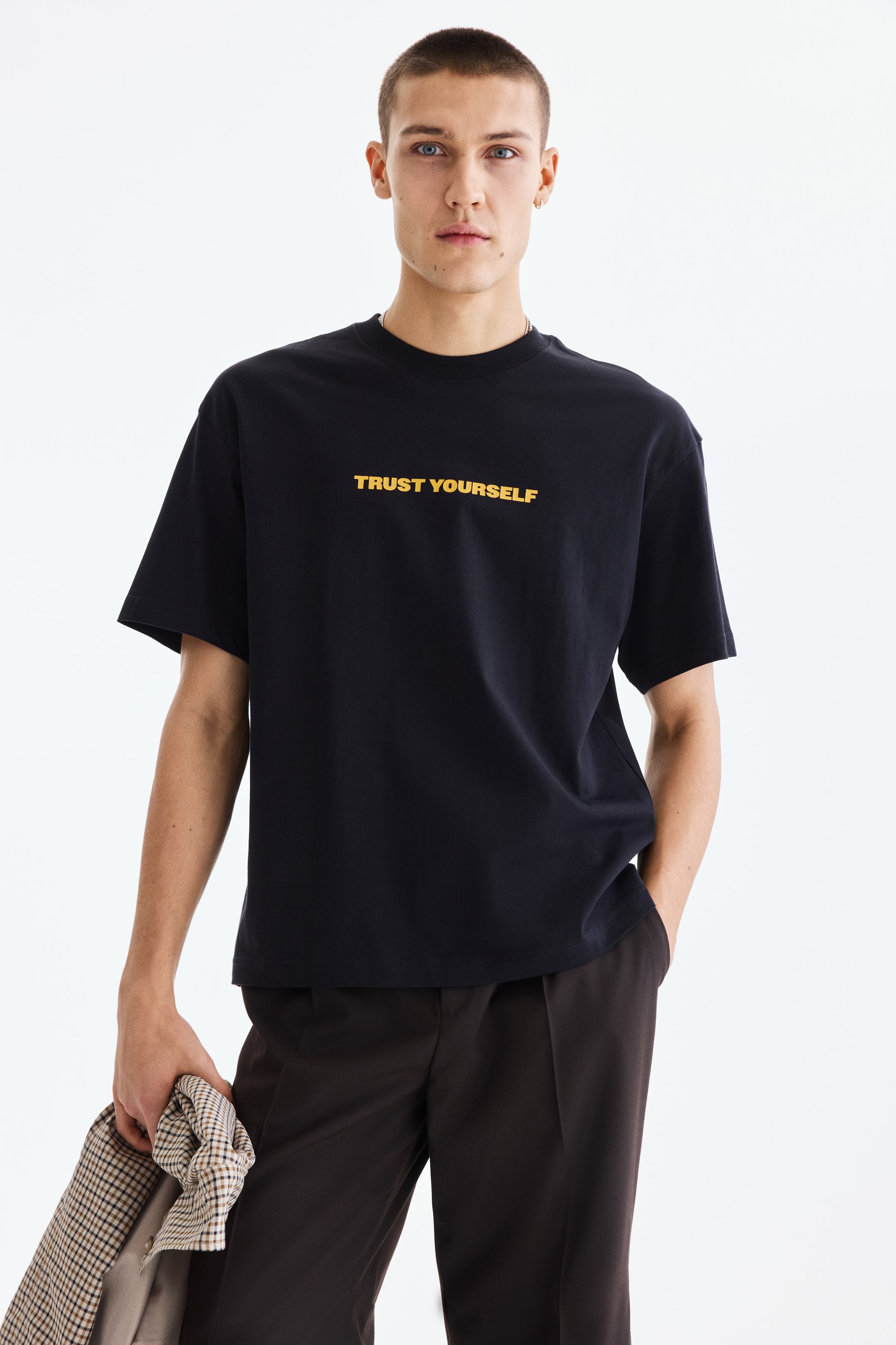 View larger image: A black crew neck t-shirt is showcased front-facing, offering a relaxed fit. It features "TRUST YOURSELF" in yellow capital letters prominently on the chest and is styled with short sleeves.