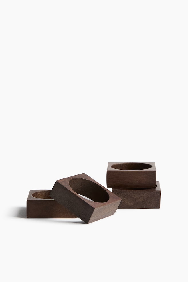 Four dark brown wooden napkin rings are displaying their square outer shapes and oval inner openings. Two rings are stacked vertically on the right, while one is leaning on its side in the center foreground, and another is partially visible behind it. - 1