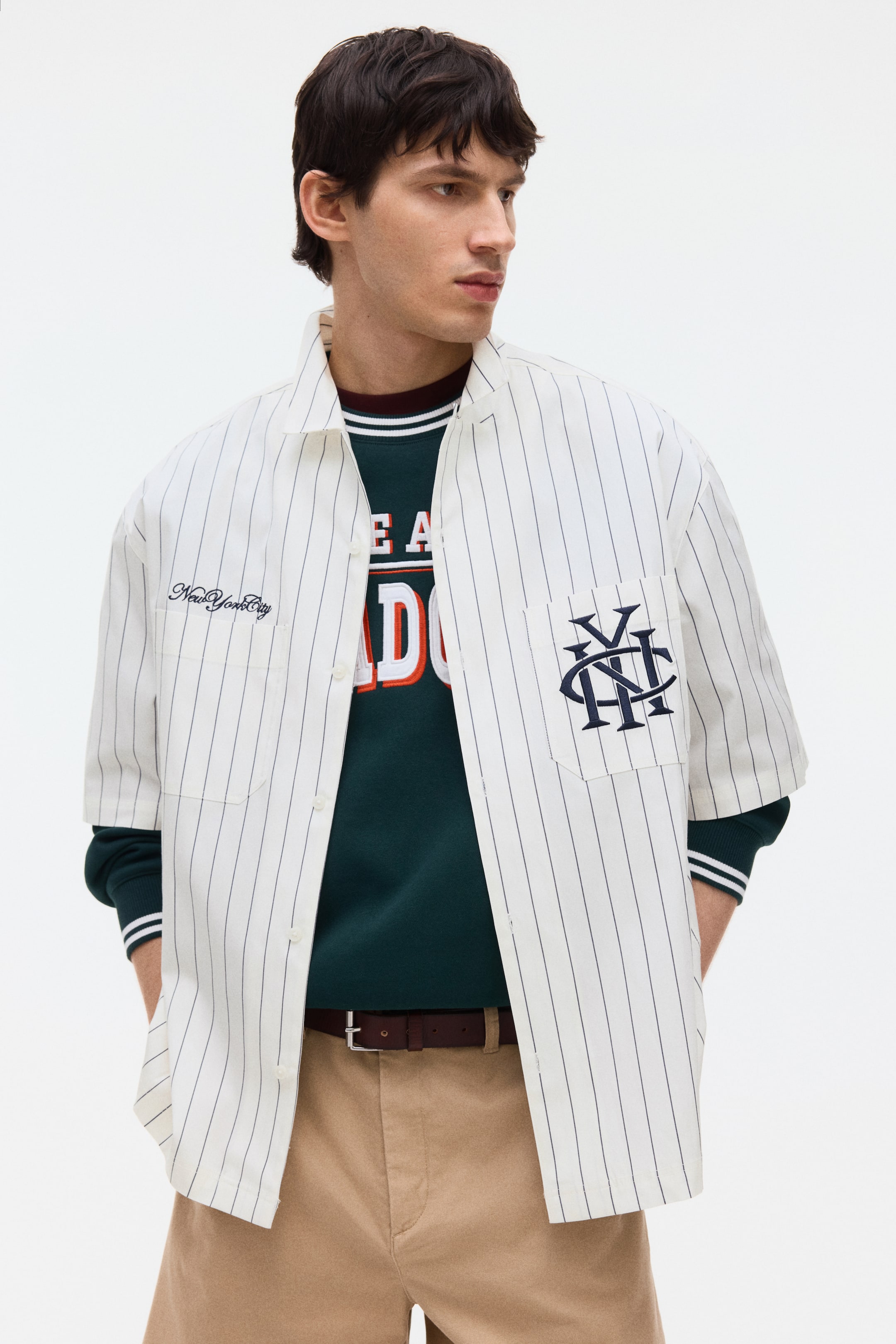 Ver imagen más grande: Una camisa blanca de manga corta con finas rayas verticales se presenta desde una vista frontal en ángulo, llevada abierta sobre una sudadera de color verde oscuro. Cuenta con un cuello extendido, cierre frontal de botones y dos bolsillos de parche en el pecho, uno bordado con "New York City" y el otro con un logotipo entrelazado "NYC".
