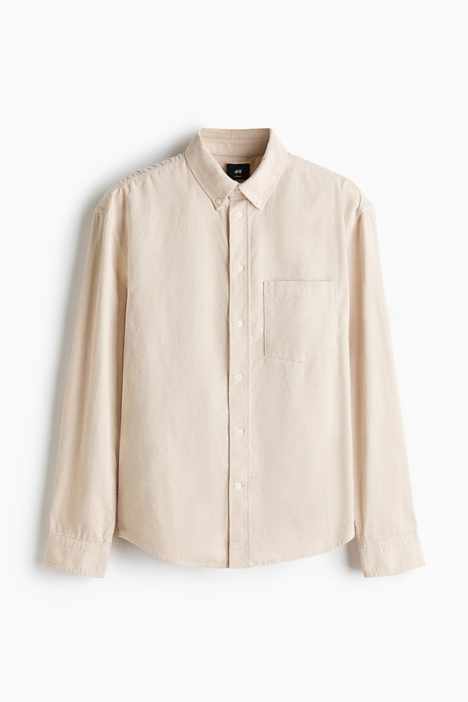 Una camisa de pana en color beige claro se presenta en vista frontal, con un cuello abotonado, una tapeta de botones central y un único bolsillo en el pecho abierto. Está diseñada con mangas largas, puños ajustables con botones, tapetas en las mangas y un dobladillo redondeado. - 1