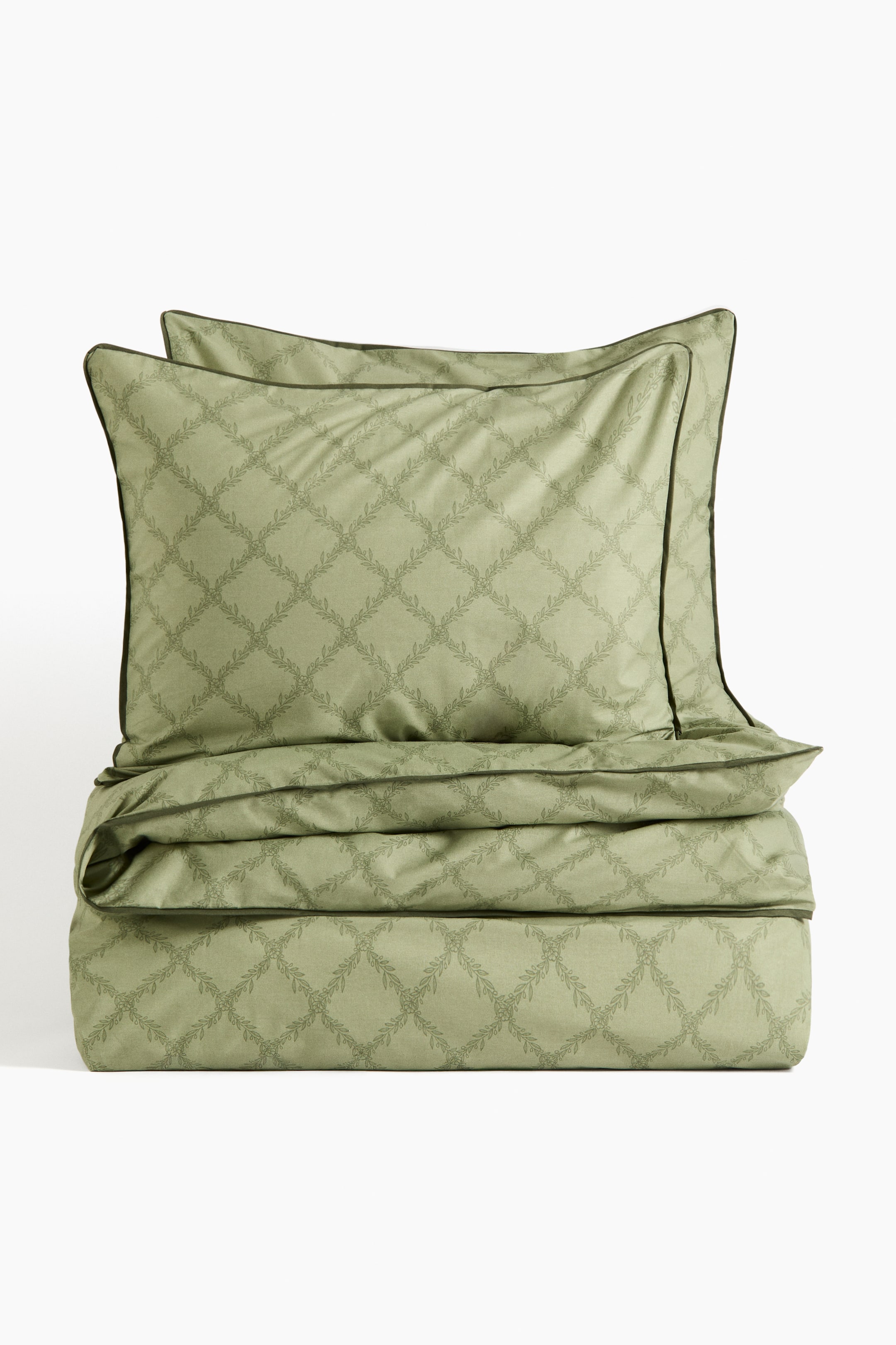 Größeres Bild ansehen: Ein hellgrünes Bettwäsche-Set präsentiert einen am unteren Ende umgeschlagenen Bettbezug und zwei übereinander gestapelte Kissenbezüge, alles von vorn gesehen. Das gesamte Set zeigt ein sich wiederholendes Gittermuster aus blättrigen Ranken und ist an den Rändern mit dunkelgrüner Paspel versehen.