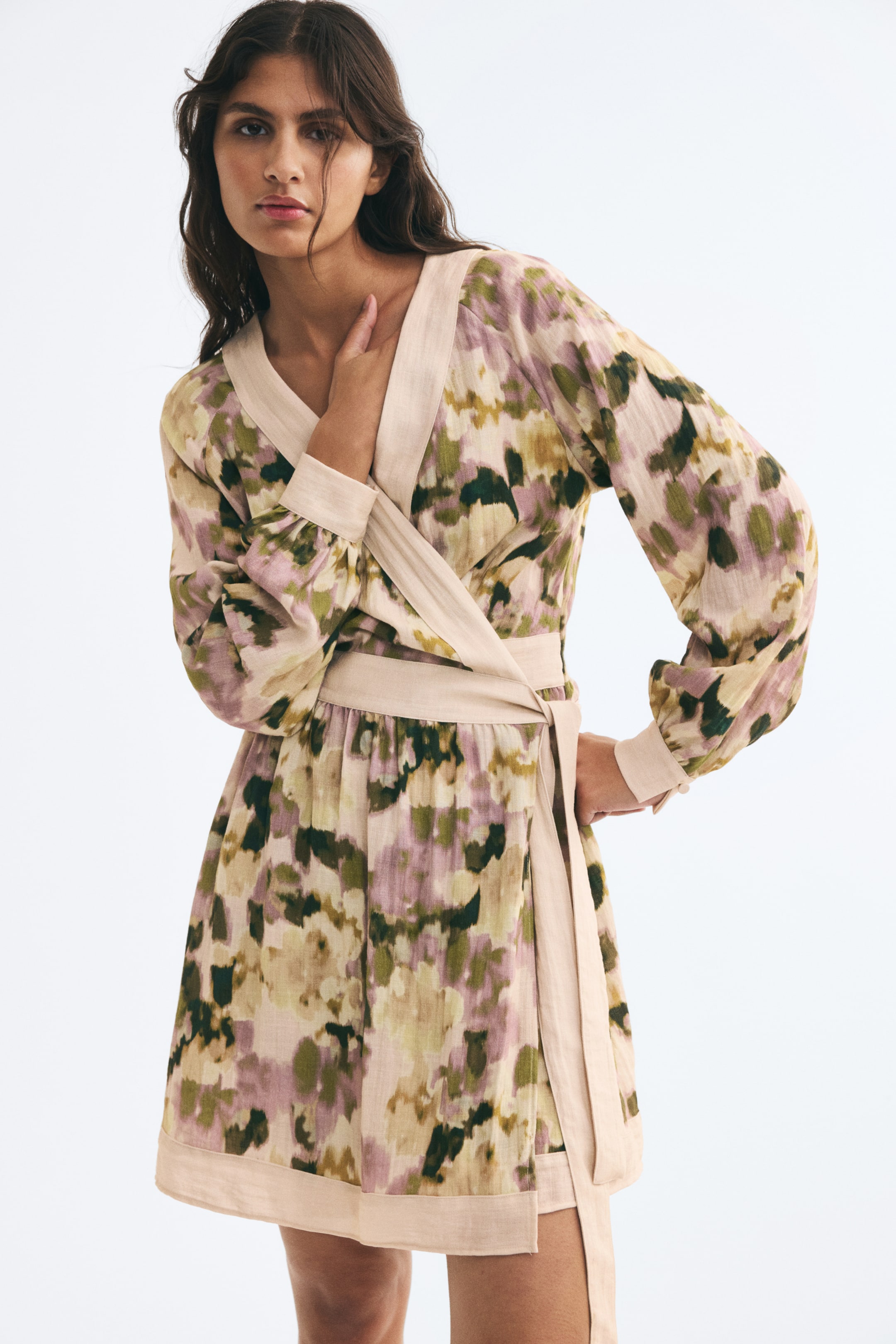 Ampliar la imagen: Un vestido corto tipo envolvente con un estampado abstracto en verde, morado y beige es usado por la modelo, quien posa con una mano cerca de la clavícula y la otra en la cadera. El vestido está diseñado con escote en V, mangas largas abullonadas y ribete beige en contraste en el escote, la cintura ajustable, los puños y el dobladillo.