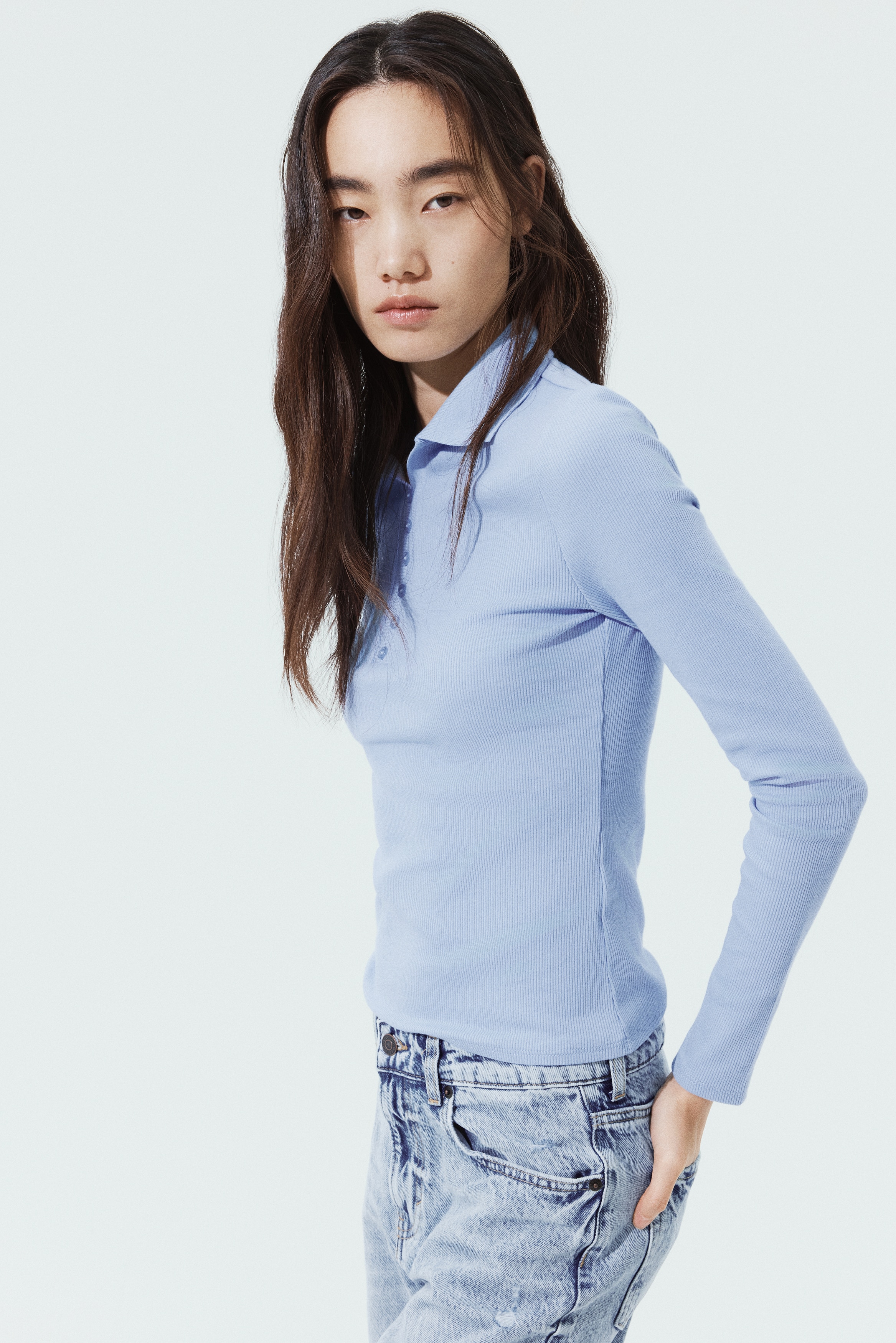 A light blue long-sleeved polo shirt displays a ribbed-knit texture and a classic collar. The shirt is shown from the front, featuring a narrow placket with six small matching buttons and a straight hem.