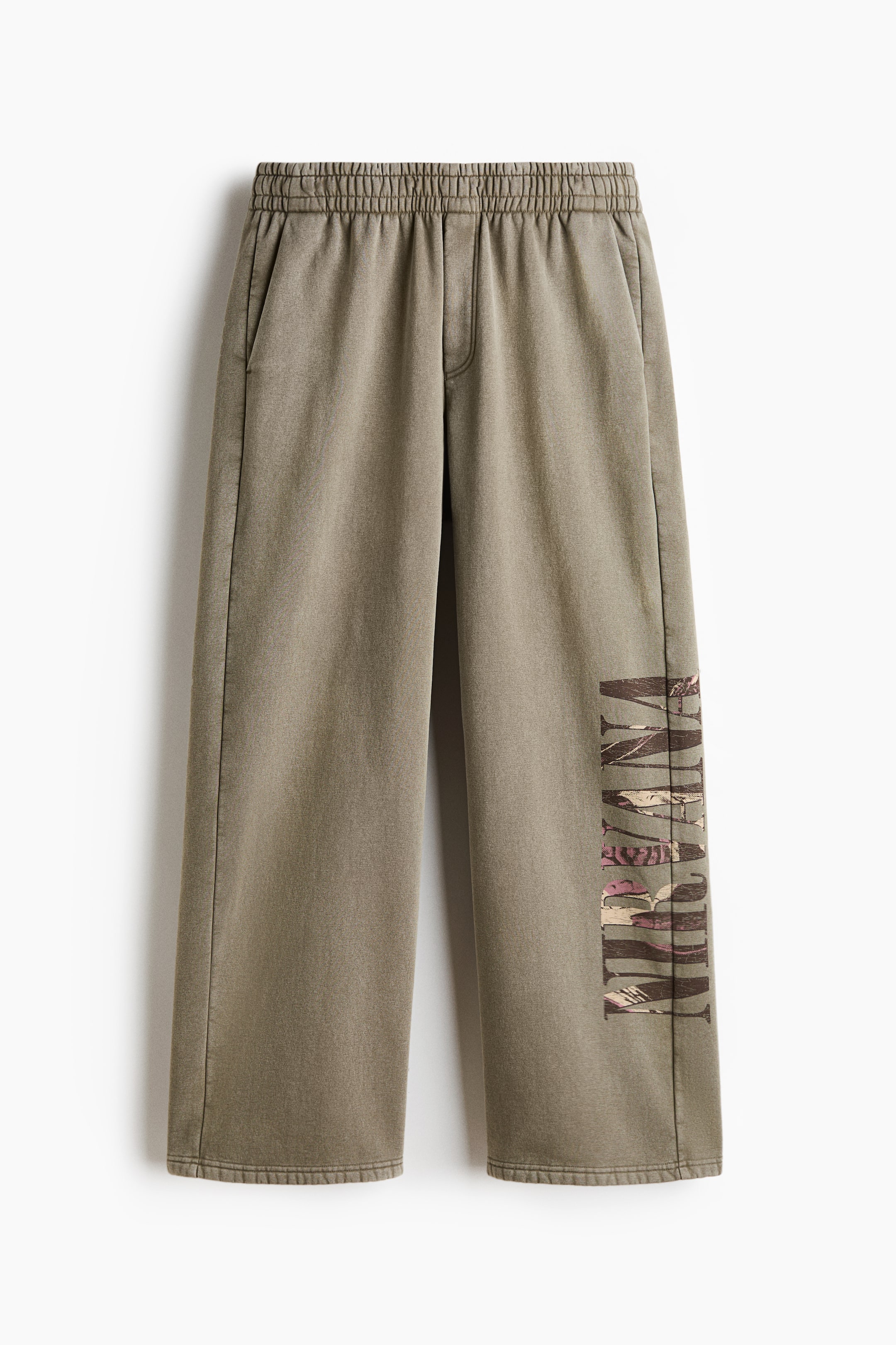 View larger image: A pair of relaxed-fit, olive green sweatpants is shown from the front, featuring an elasticated waistband and side slip pockets. The lower left leg displays a vertical graphic print of the word "NIRVANA" with a pink and brown patterned fill.