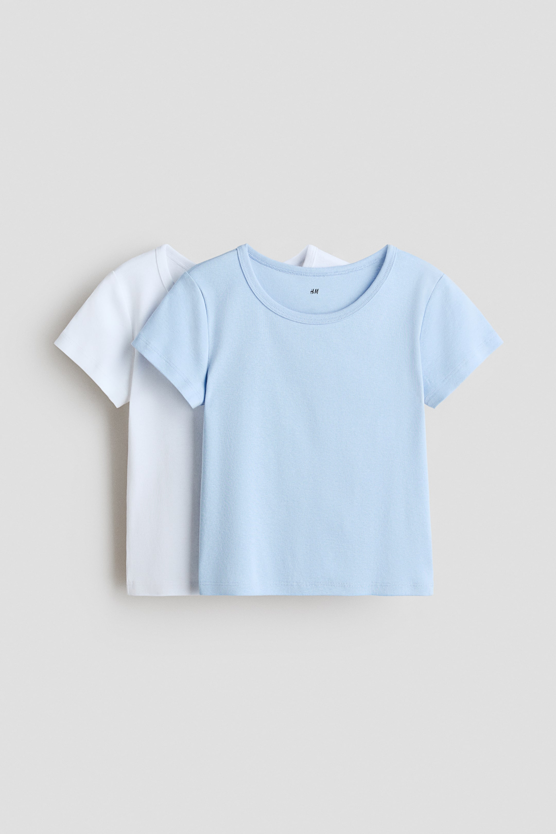 View larger image: Two short-sleeved t-shirts are layered, with a light blue t-shirt in the FRONT partially obscuring a white t-shirt. Both t-shirts feature round necklines, and the light blue t-shirt displays a visible 'H&M' label at the neck.
