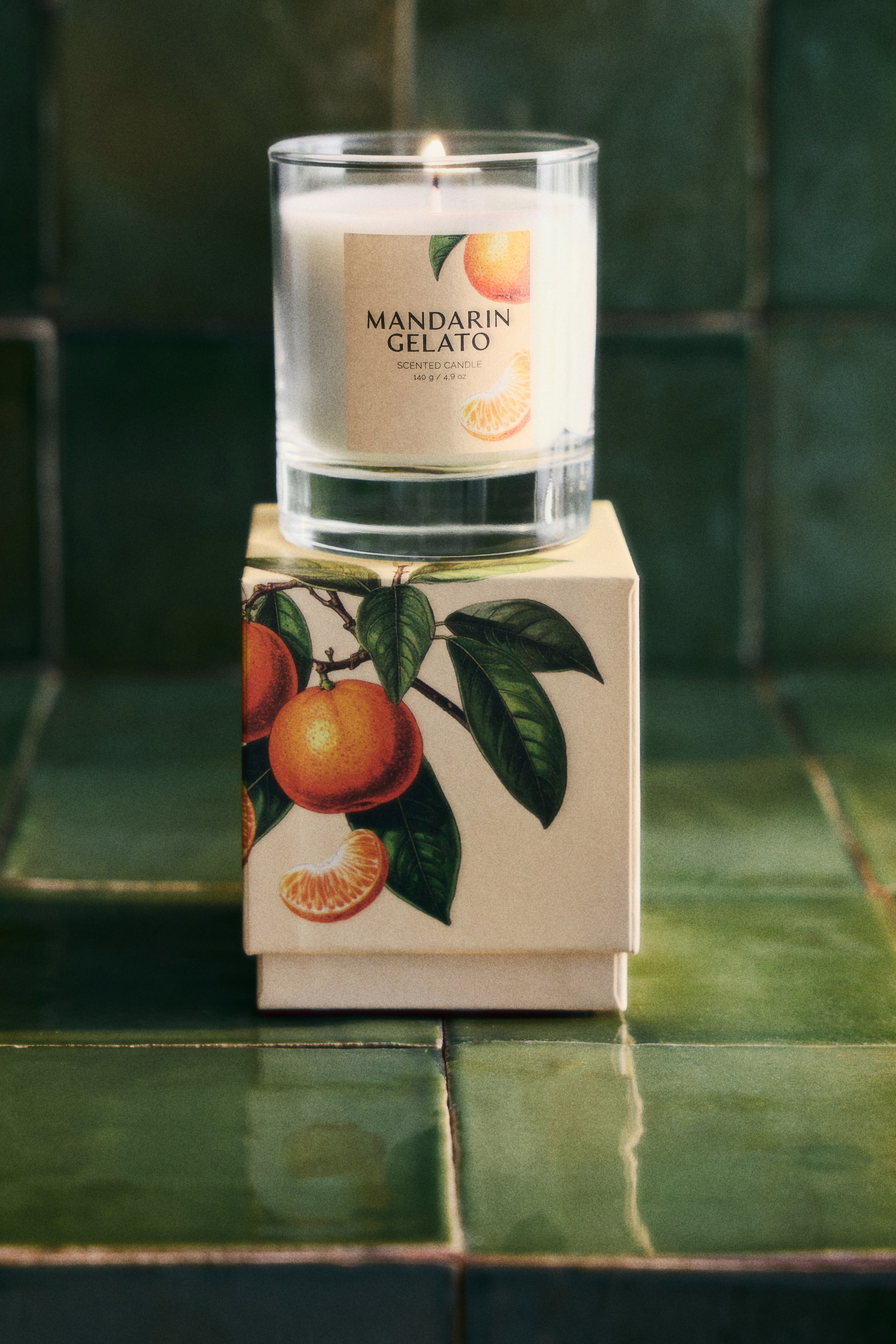 View larger image: A scented candle in a clear glass container with a thick base is perched atop its decorative gift box. The candle's front label showcases 'MANDARIN GELATO' alongside mandarin fruit illustrations, while the gift box is adorned with patterns of mandarin branches and fruit.