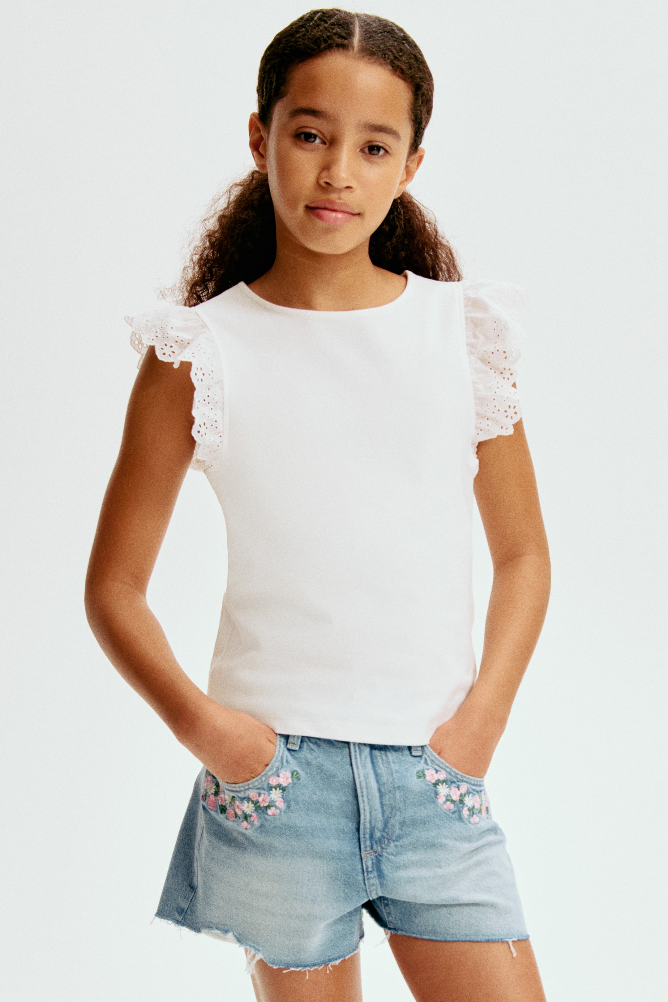 View larger image: A white top featuring a round neckline and short ruffled sleeves with eyelet detailing. Light wash denim shorts showcasing a raw hem and floral embroidery on the front pockets.