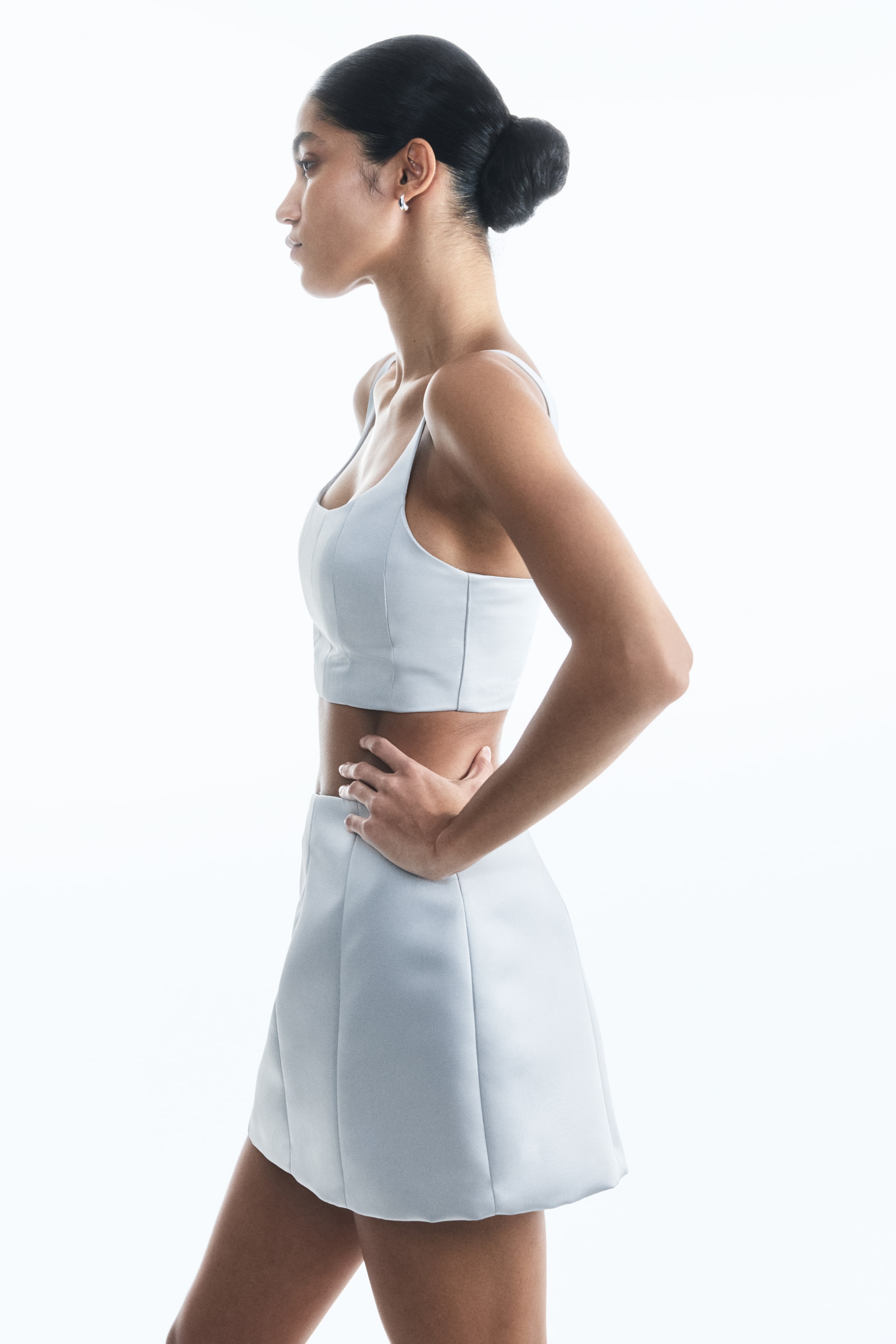 View larger image: A light blue two-piece set is being presented from a side profile. The set is featuring a paneled crop top with thin straps and a structured A-line mini skirt, both made from a smooth, structured fabric.