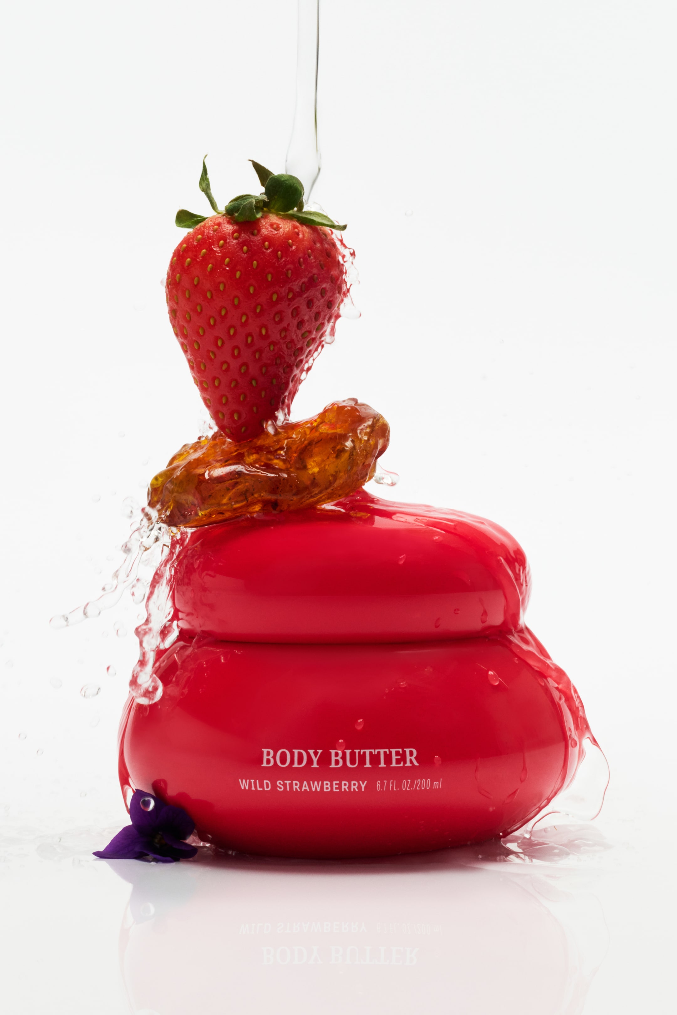 Agrandir l'image: Un pot de beurre corporel rouge est orienté vers l'avant, affichant son étiquette "BEURRE CORPOREL FRAISE SAUVAGE 6.7 FL. OZ. / 200 ml" sur sa face avant. Une fraise repose sur un objet de couleur ambre placé sur le couvercle du pot, tandis que de l'eau coule dessus et éclabousse les côtés du pot.