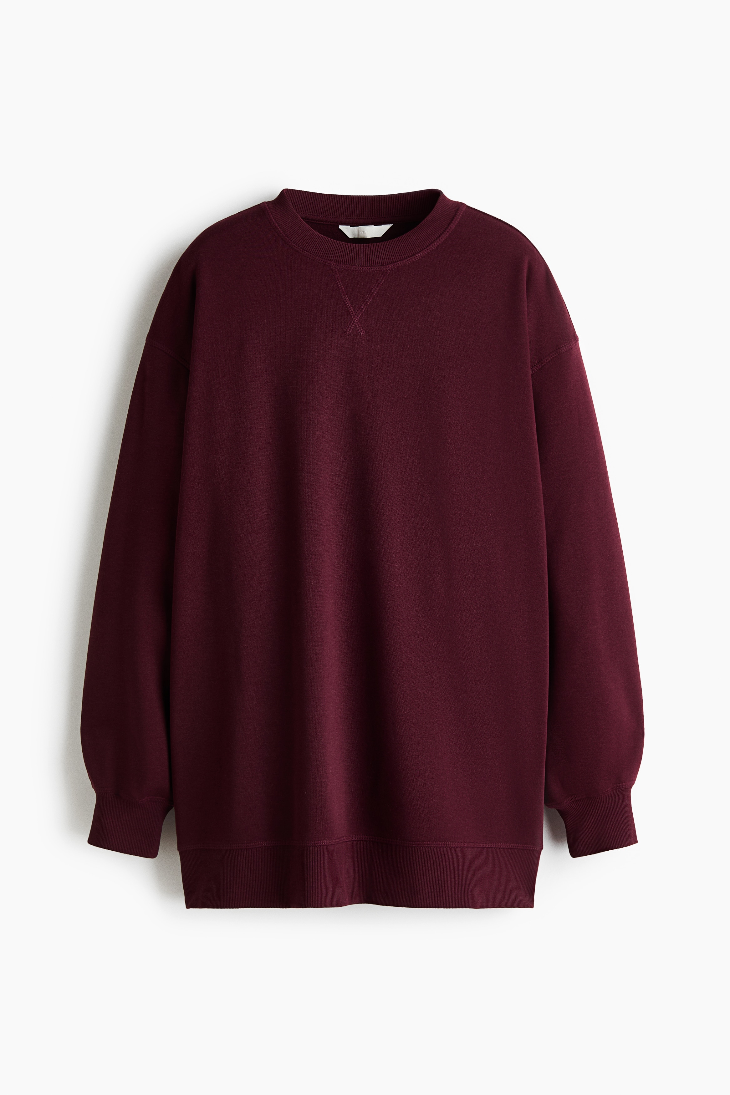 Model ma na sobie oversize’ową bordową bluzę z obniżoną linią ramion, długimi rękawami zakończonymi prążkowanymi mankietami oraz prążkowanym, okrągłym dekoltem. Bluza jest zestawiona z czarnymi legginsami.