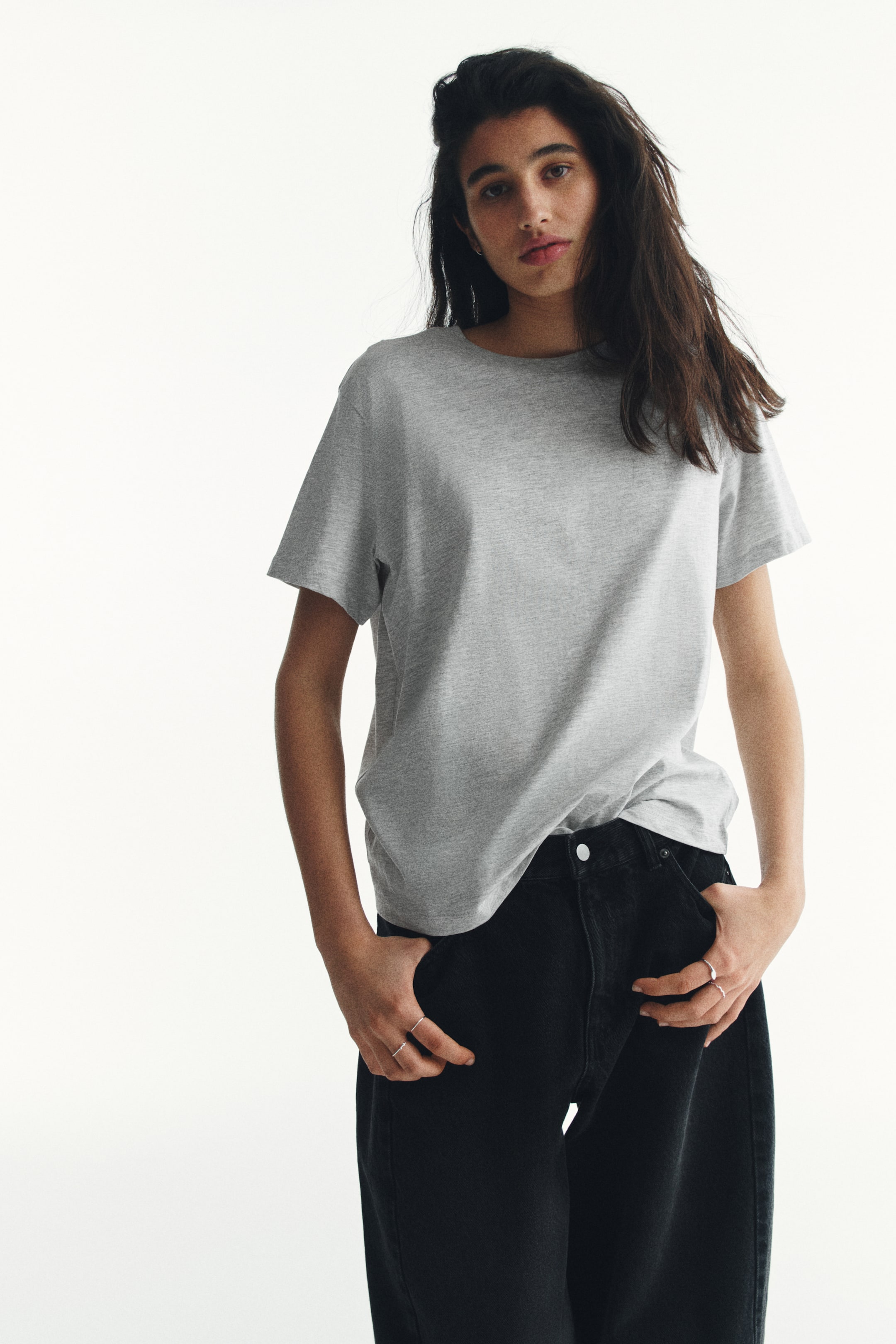Vis større bilde: En lys grå t-skjorte med korte ermer og ribbet rund hals bæres, lett stukket inn i sorte, høytlivs jeans med sølvfarget knappelukking.