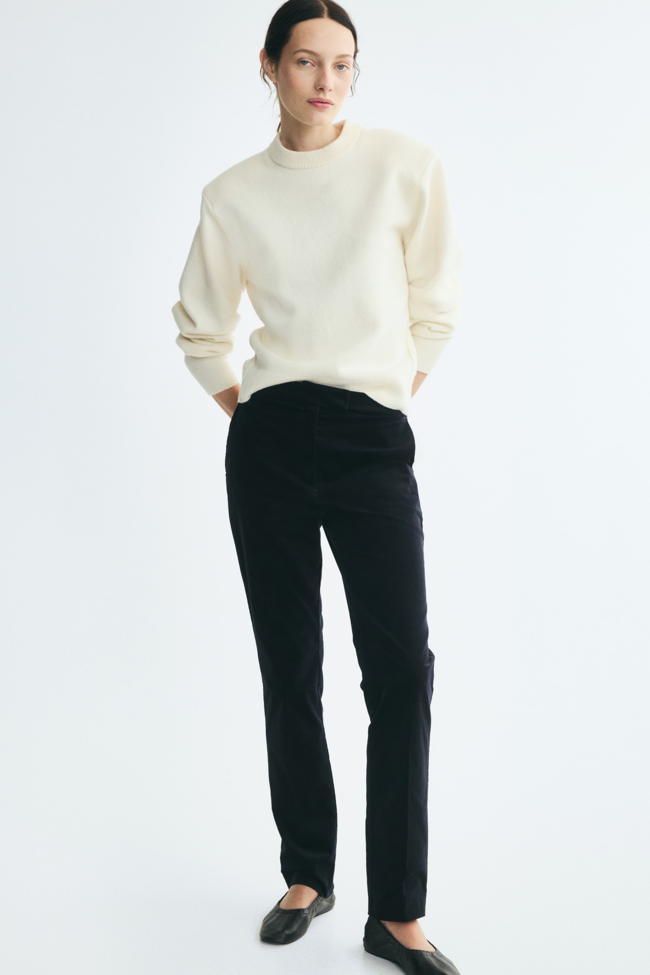 View larger image: The model is wearing fitted black velvet pants with slim legs and front creases. The pants are styled with a cream crewneck sweater and black leather flats.
