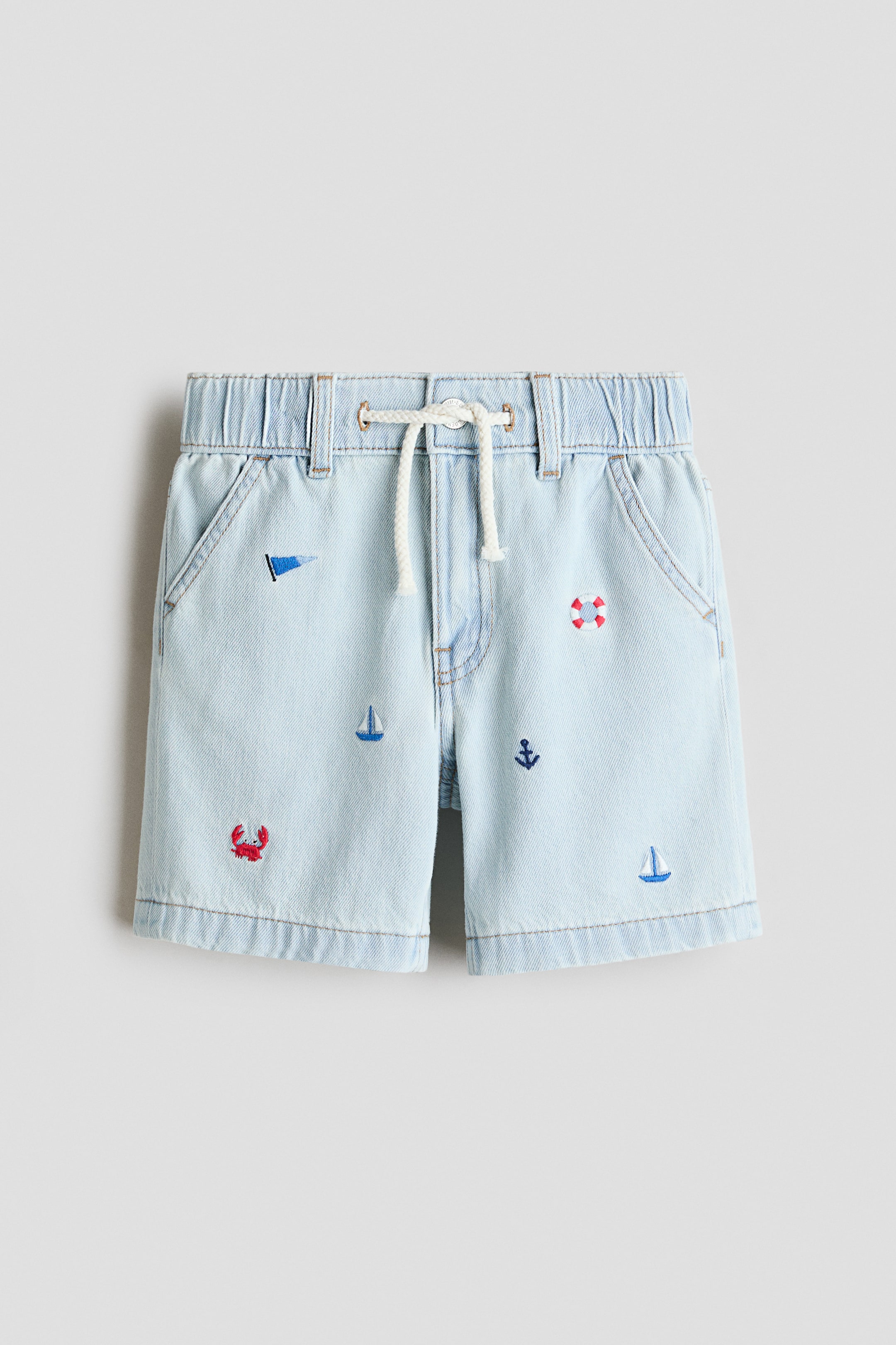 View larger image: Denim 短褲從正面展示，採用淺藍色水洗設計，並配有彈性腰帶，中央綁有白色繩索抽繩。斜插前袋和各種海洋主題刺繡，包括帆船、螃蟹、錨、旗幟和救生圈，為短褲增添特色。