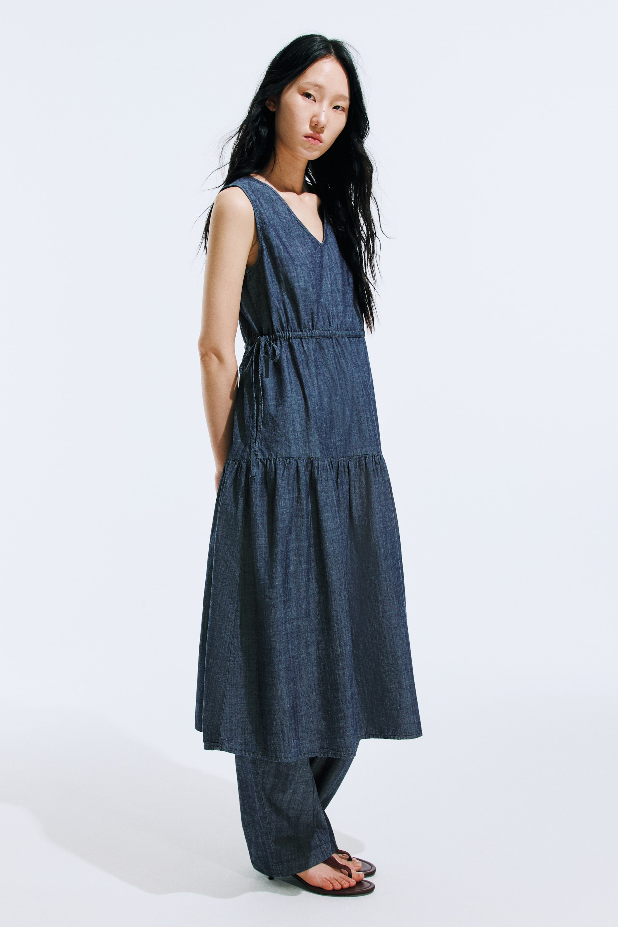 Xem ảnh lớn hơn: Một chiếc váy denim cotton không tay dài đến đầu gối, có cổ chữ V, dây rút hai đầu ở eo và một tầng xếp ly trên hông, đang được người mẫu mặc, người đang phối nó với quần denim.