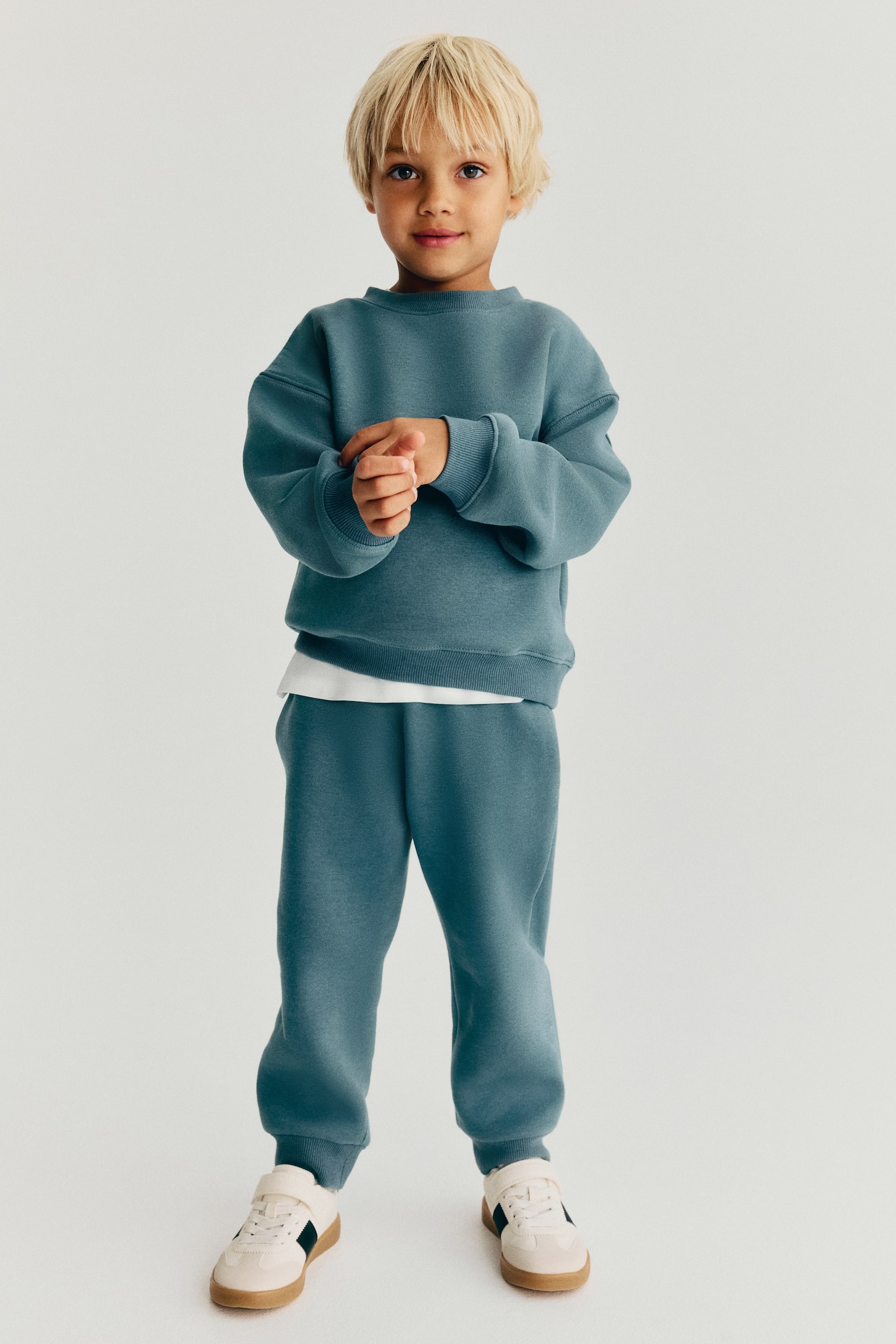 A front view of relaxed-fit track pants in a blue-grey sweat fabric, featuring ribbed cuffs at the ankles. The track pants are styled as a set with a matching crewneck sweatshirt and cream-coloured sneakers. - 1