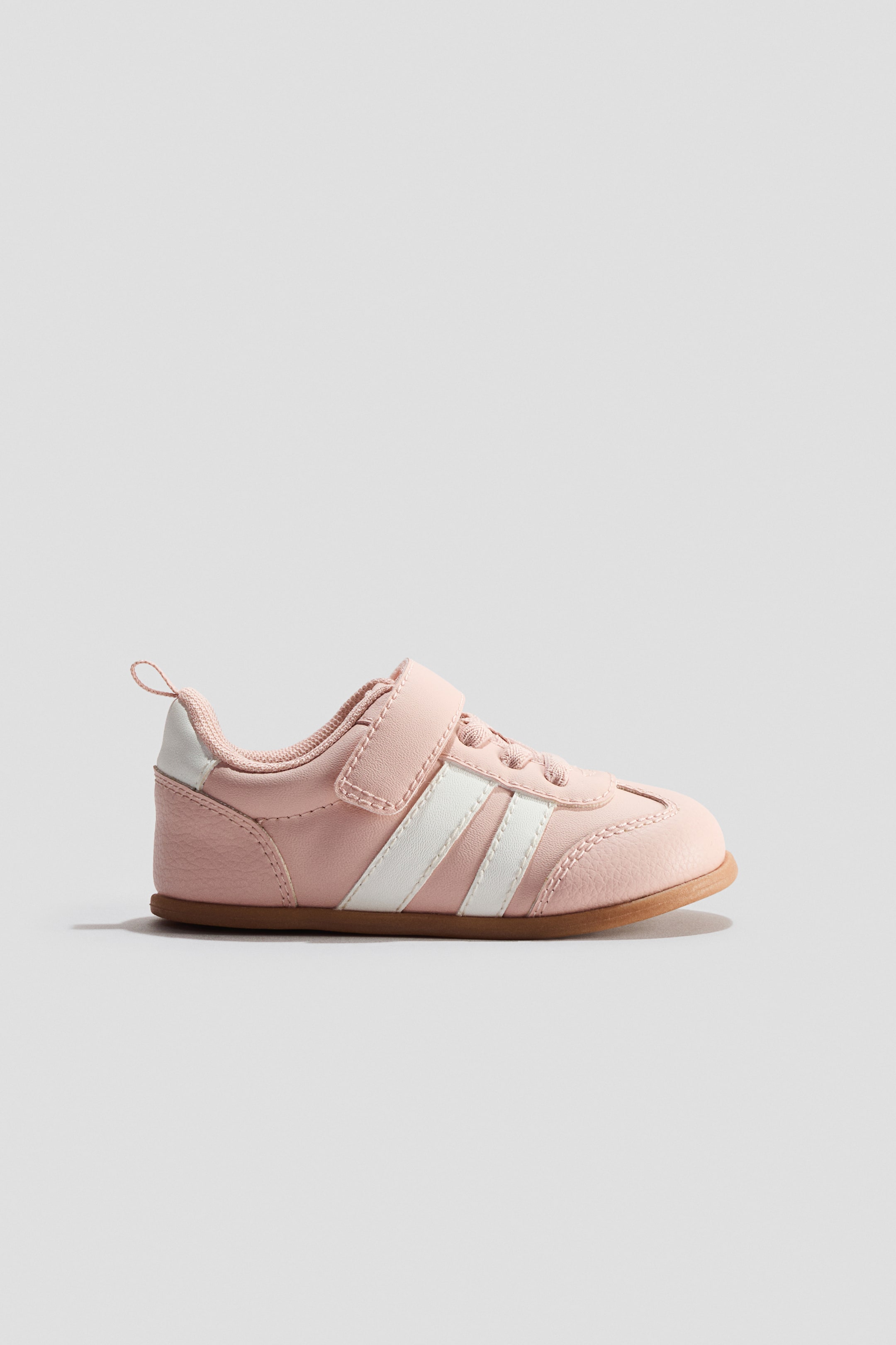 View larger image: The pink trainer showcases two white stripes along its side, complemented by a hook-and-loop strap and faux laces. It also features a white heel accent, a rear pull tab, and a brown sole.