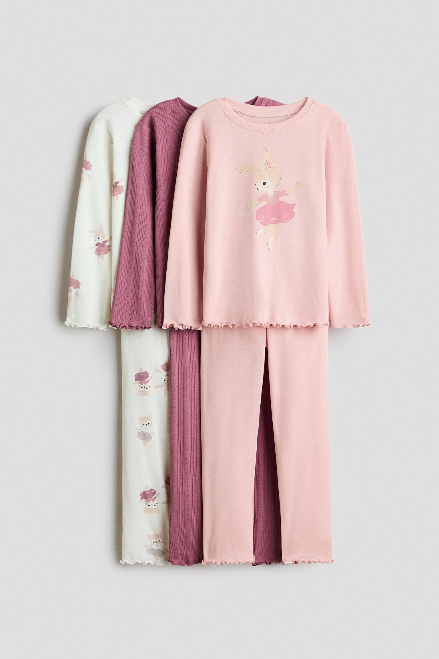 ÃÃ§ adet pamuklu jarse pijama seti, Ã¶nden bakacak Åekilde birlikte asÄ±lÄ± duruyor; uzun kollu Ã¼stler ve overloklu marul kenar paÃ§alÄ± uyumlu pantolonlar iÃ§eriyor. Ãndeki set, balerin tavÅan grafik baskÄ±lÄ± aÃ§Ä±k pembe renkte; ortadaki set, nervÃ¼rlÃ¼ Ã¶rme dokuda dÃ¼z koyu gÃ¼l kurusu; arkadaki set ise her yerine uygulanmÄ±Å tavÅan desenli krem beyaz renkte.